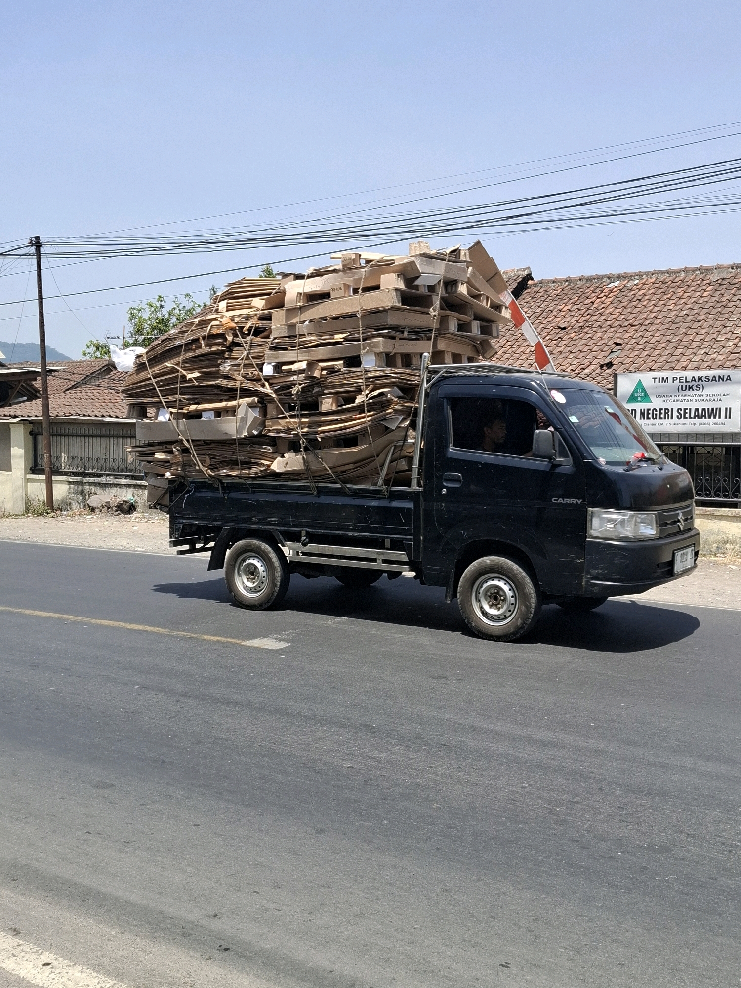 A black Suzuki Carry pickup truck is driving on a paved road in Selaawi, Indonesia. The truck bed is heavily loaded with a large stack of flattened cardboard boxes and wooden pallets, secured with ropes. The boxes are piled high, exceeding the height of the truck's cab. A person is visible inside the driver's side of the cab. In the background, there are buildings with red tile roofs and a sign that reads "TIM PELAKSANA (UKS) USAHA KESEHATAN SEKOLAH KECAMATAN SUKARAJA SDN NEGERI SELAAWI II". The sky is clear and blue. The scene appears to be a sunny day.