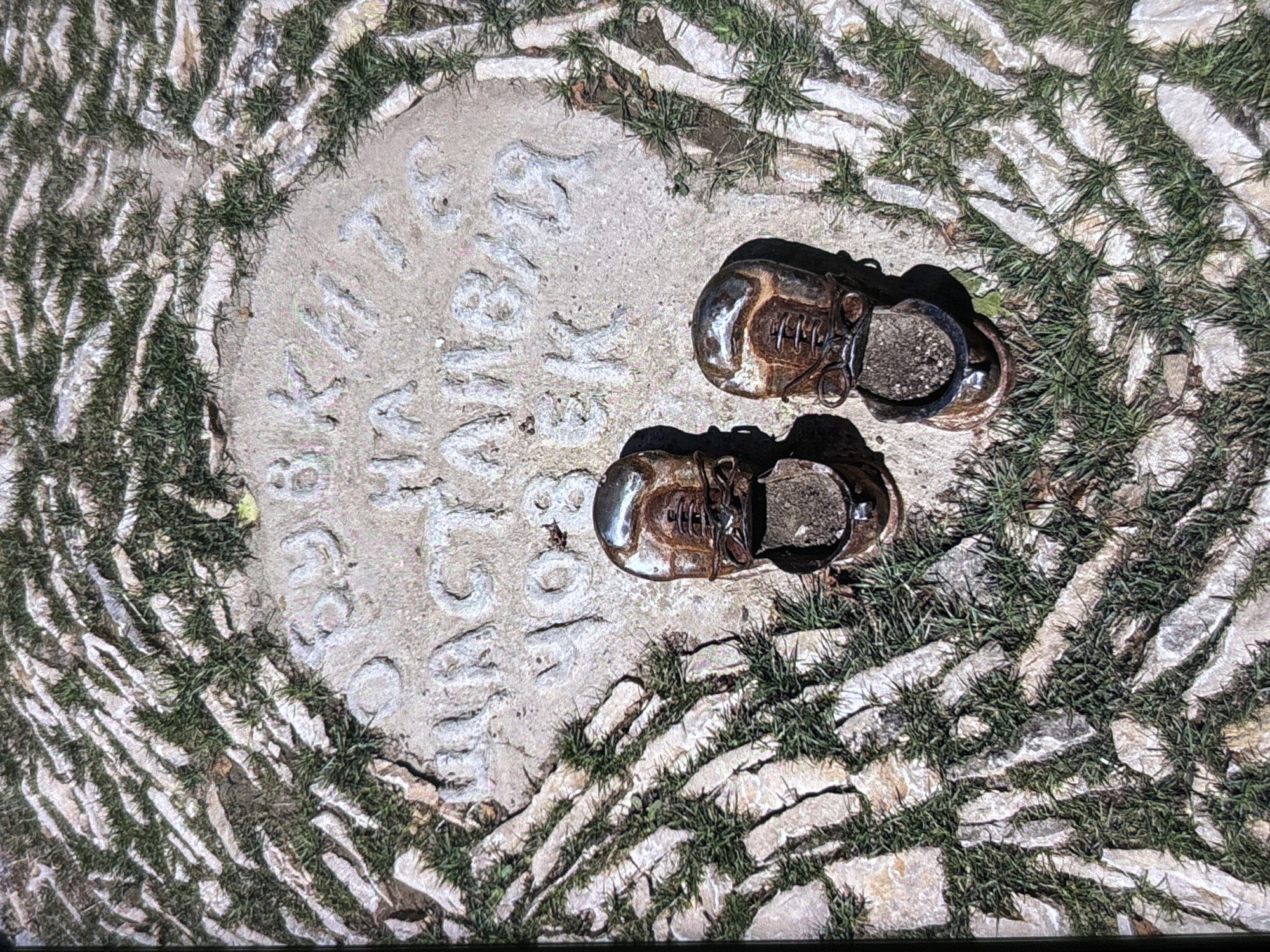 An overhead shot captures two weathered, miniature shoes, likely made of patinated metal such as bronze or copper, resting on a circular concrete plaque. The plaque is set into a ground surface composed of irregular, light-colored flagstones with vibrant green grass growing between them, radiating outwards in a dynamic pattern.
The shoes, resembling old, well-worn boots with visible laces and eyelets, are positioned on the right side of the plaque. They cast sharp shadows, indicating bright, direct sunlight. The left shoe is slightly higher than the right, and their interiors appear filled with soil or small pebbles.
Embossed or carved into the concrete plaque is text arranged in several lines. The top line reads "THE SPIRIT OF", followed by the prominent central line "WOLVERHAMPTON". Below this, the text continues with "MARKET TRADERS". At the bottom of the plaque, in smaller lettering, are the years "1880" and "1994".
The scene suggests an outdoor memorial or tribute, commemorating the market traders of Wolverhampton for the period spanning 1880 to 1994. The worn shoes likely symbolize the journey and hard work associated with their profession. The setting is bright and sunny, conveying a sense of historical commemoration in a public space within Wolverhampton, United Kingdom.