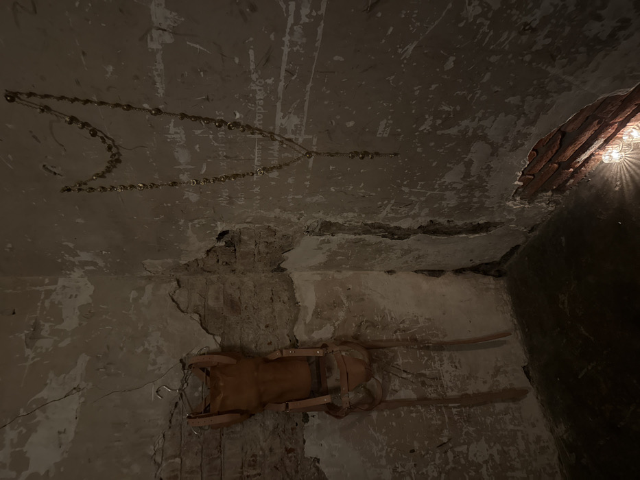 An eerie interior scene shows a dilapidated room with crumbling walls and ceiling. A string of lights hangs from the ceiling, forming a decorative pattern. Below, a vintage piece of furniture, possibly a saddle or chair, is mounted on the wall. The room's ambiance is dim, with a light source visible on the right, casting shadows and emphasizing the room's aged, decaying condition.