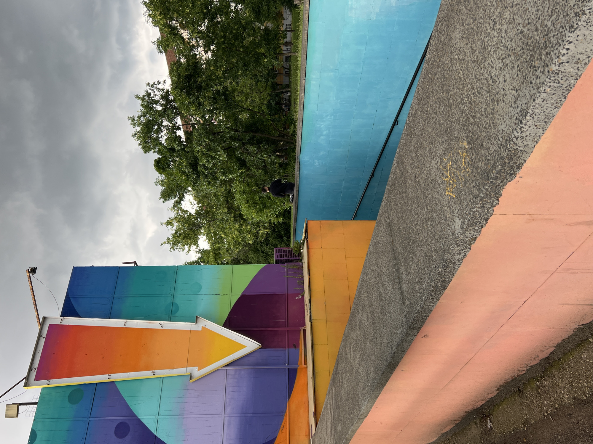 The image, rotated 90 degrees clockwise, presents a vibrant outdoor scene characterized by diverse surfaces and strong colors under an overcast sky. In the lower left, a large wall is adorned with a colorful, geometric design featuring shades of blue, green, and purple. A prominent, reflective arrow sign in bright orange and yellow is mounted on this wall, pointing to the left. Its metal frame shows signs of age and rust, with a light fixture visible above it.
Towards the upper middle of the frame, a person in dark attire stands near a bright blue-tiled wall, which stretches horizontally across the midground. A black handrail runs along the top of this blue wall. Behind it, and filling the upper left section of the image, are dense green trees under a dark, cloudy sky, partially obscuring distant buildings.
On the right side, a diagonally oriented, coarse dark gray concrete surface features visible aggregate and faint, illegible yellow markings or graffiti. Below and adjacent to this, a lighter peach-colored concrete surface also runs diagonally, showing signs of wear and a textured, slightly uneven edge with hints of dirt and moss at its base. Below the blue wall and positioned to the left of the gray concrete, a smaller, yellow-orange wall is visible. The scene captures an urban landscape combining street art, architectural elements, and natural greenery under a diffuse daylight sky, consistent with a location such as Sofia, Bulgaria.