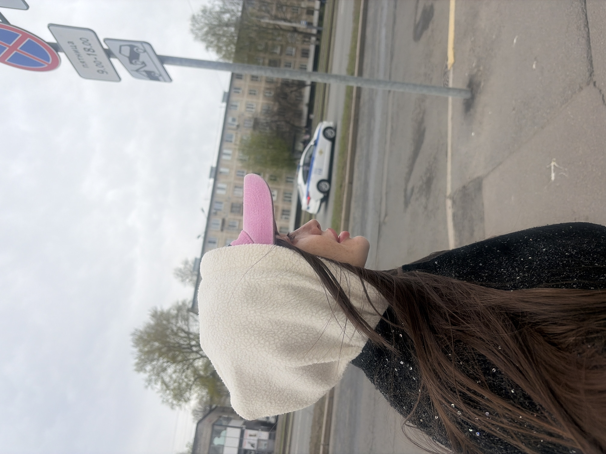 A medium, sideways shot focuses on a person wearing a white, furry bucket hat with pink ears, gazing upward. The person has brown hair and is wearing a dark, sequined garment. In the background is a typical Saint Petersburg street scene featuring a light-colored building, trees, and a road with a white-and-blue car. A traffic sign is also visible in the upper part of the background.