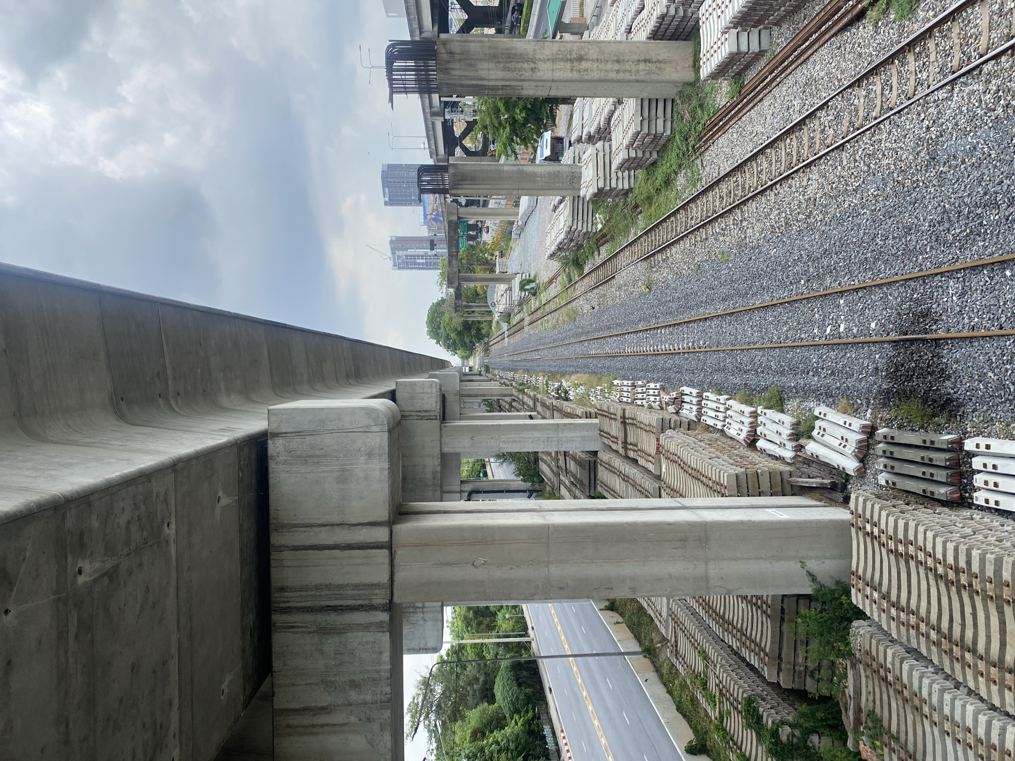 The image depicts an elevated view along a stretch of railway tracks and infrastructure in Bangkok, Thailand. The scene includes a railway line with gravel ballast, stacks of precast concrete elements, a concrete elevated structure to the left, and a road visible beneath the elevated structures. Tall buildings can be seen in the distance. The sky is overcast.