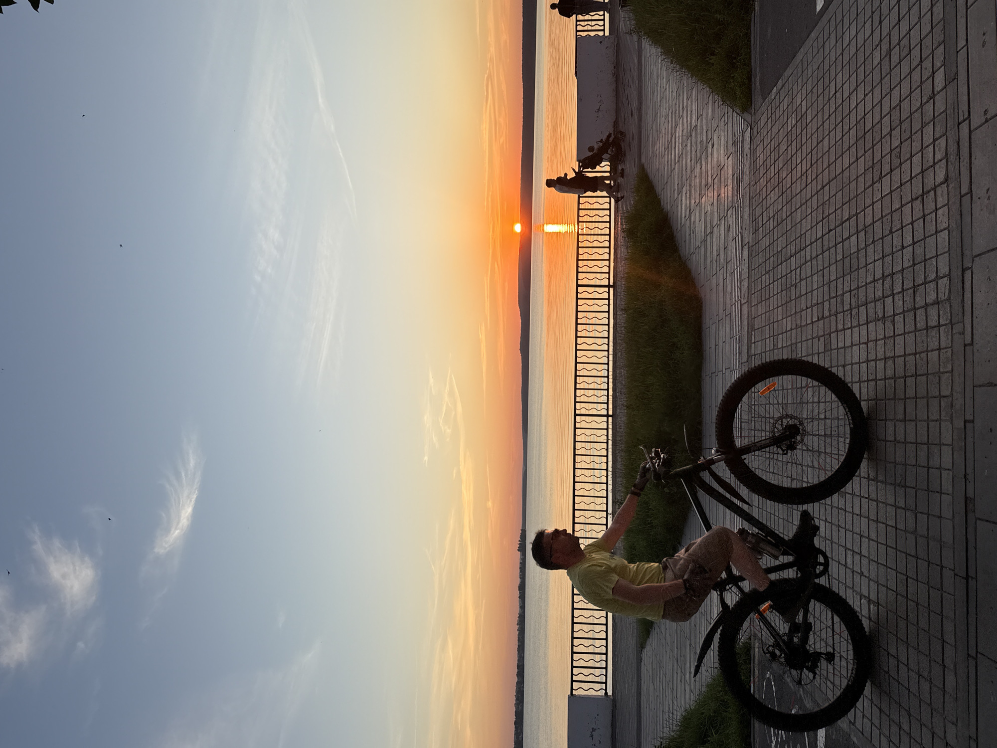 A full-shot vertical image captures a man cycling on a paved path by a body of water during sunset. The man, wearing glasses, a yellow t-shirt, and shorts, is positioned in the lower-left quadrant of the frame, riding a black mountain bike. He is facing left, with his right hand on the handlebars. The path is paved with square tiles and is bordered by green grass on the left.
In the background, a black decorative fence runs along the edge of the water. Beyond the fence, a vast lake stretches to the horizon, reflecting the vibrant orange and yellow hues of the setting sun. The sky above is a gradient of blue and orange, with scattered clouds and two small birds flying.
Further in the background, on the far side of the lake, a treeline is visible. On the paved path to the right of the cyclist, silhouettes of other people can be seen walking, including a person pushing a stroller. The overall atmosphere is peaceful and scenic, indicative of an evening stroll or recreational activity in a park or promenade setting. The location is Votkinsk, Russia, a city known for its picturesque waterfront.