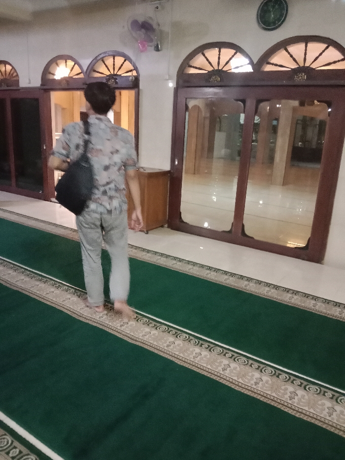 The scene is inside a mosque located at -6.976, 110.331 in Tugu, Indonesia. A single man, wearing a light-colored patterned shirt and pants, walks barefoot on a green patterned prayer rug. He carries a black bag over his left shoulder. There is an open door with a window to his right. The door leads to another room with tiled floors, and the window is visible from outside. There is a wooden cabinet next to the open door and a ceiling fan. The floor is tiled and white. There is a clock on the wall to the right of the door. The walls are white.