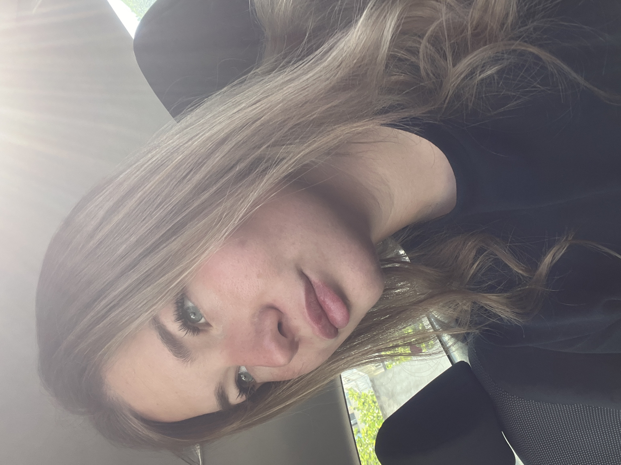 A young woman with light blonde hair, fair skin, and blue eyes is posing for a photo. She is wearing a dark-colored top. The background suggests she is in a vehicle, possibly a car. Through the window, there are glimpses of a building and greenery. The location might be Budva, Montenegro, based on the general architectural style and vegetation visible in the background. However, without more information, confirming the location definitively is impossible.