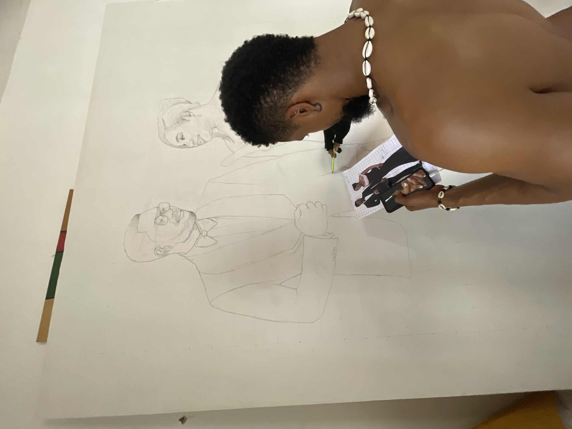 A man is drawing on a large canvas in what appears to be a room in Ado, Nigeria. The canvas shows a pencil sketch of two people, one in a suit and tie and another person above and slightly behind them. The man holds a reference photo in his left hand. He is using a pencil to create a drawing on the canvas. The man is wearing a necklace of white shells and a matching bracelet. The room is sparsely decorated. The man is shirtless and appears to be sitting or kneeling in front of the canvas.