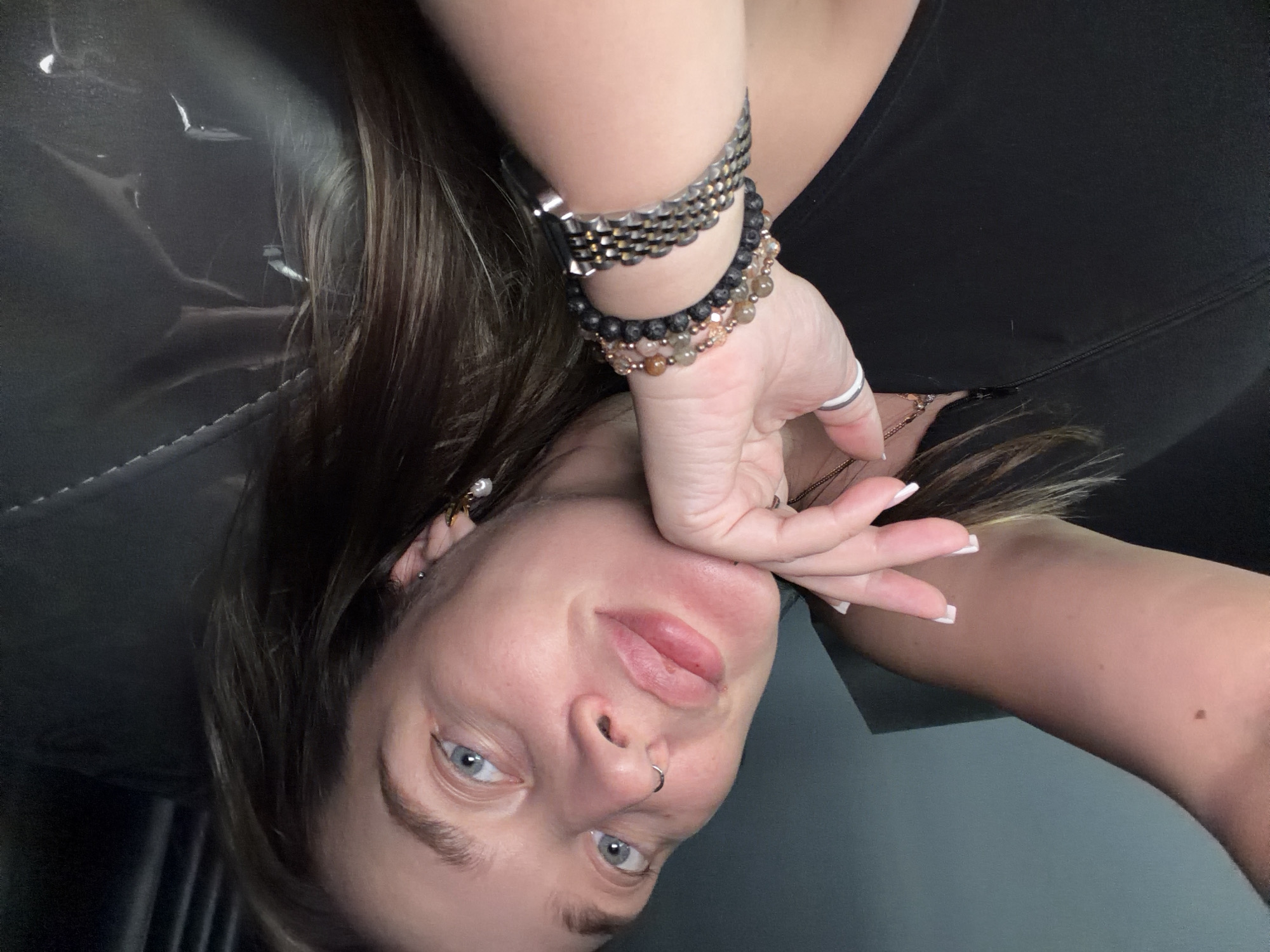 A woman with fair skin, blue eyes, and brown hair poses upside down, wearing multiple bracelets on her left wrist. Her nails are painted white with a square shape. She is wearing a black top, and a nose ring is visible. The background appears to be a dark, possibly indoor, setting. The location is not discernible as Moscow, Russia.