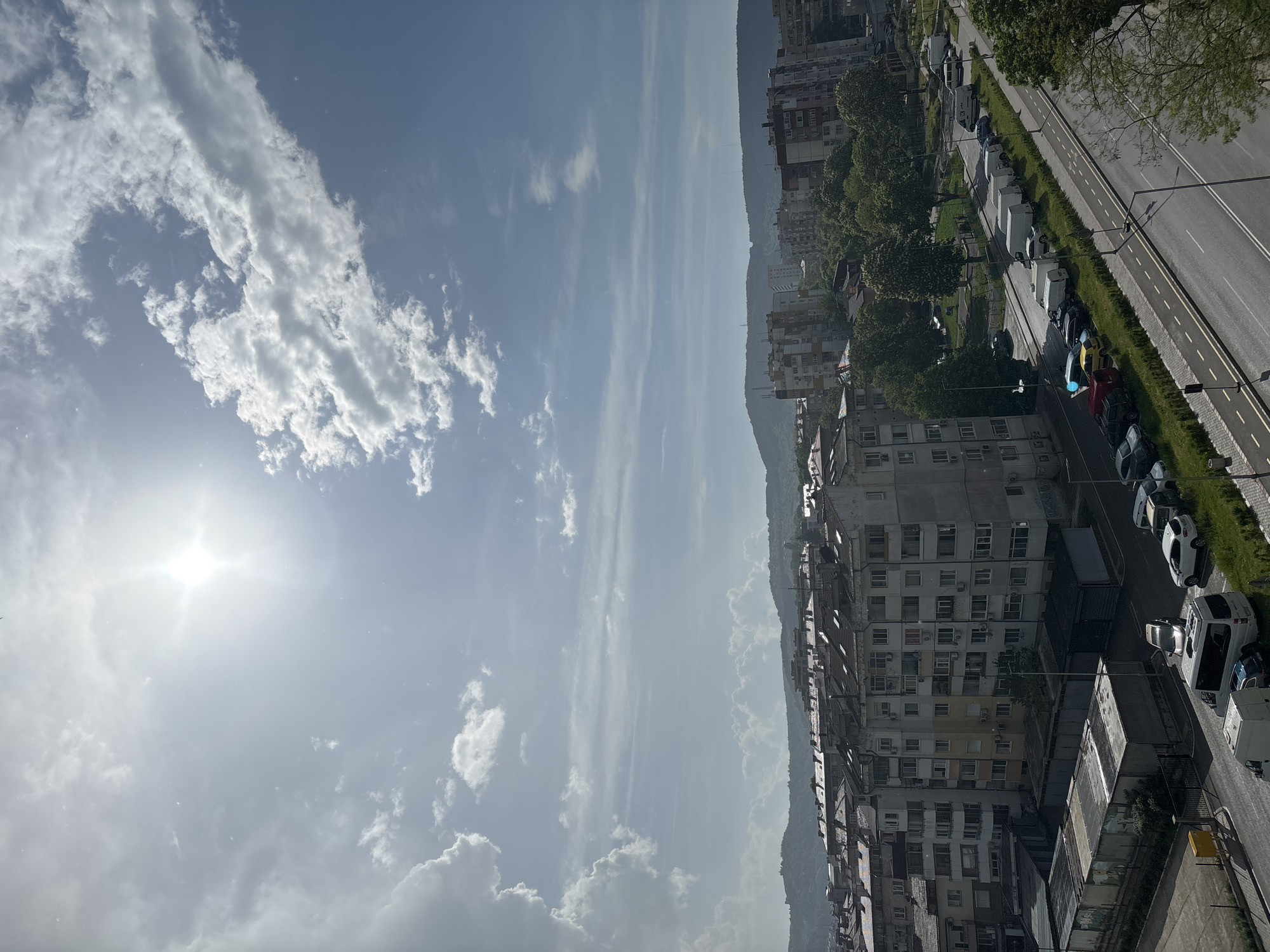 The image shows a cityscape in an unknown location, with a view from an elevated position, possibly a building. The sky is predominantly blue with scattered white clouds and the sun shining brightly. Below the sky is a mountain range, and at the bottom of the image are densely packed buildings that appear to be residential, along with a road lined with parked cars and some trees. The orientation of the image is such that the sky is on the left side, and the cityscape is on the right.
