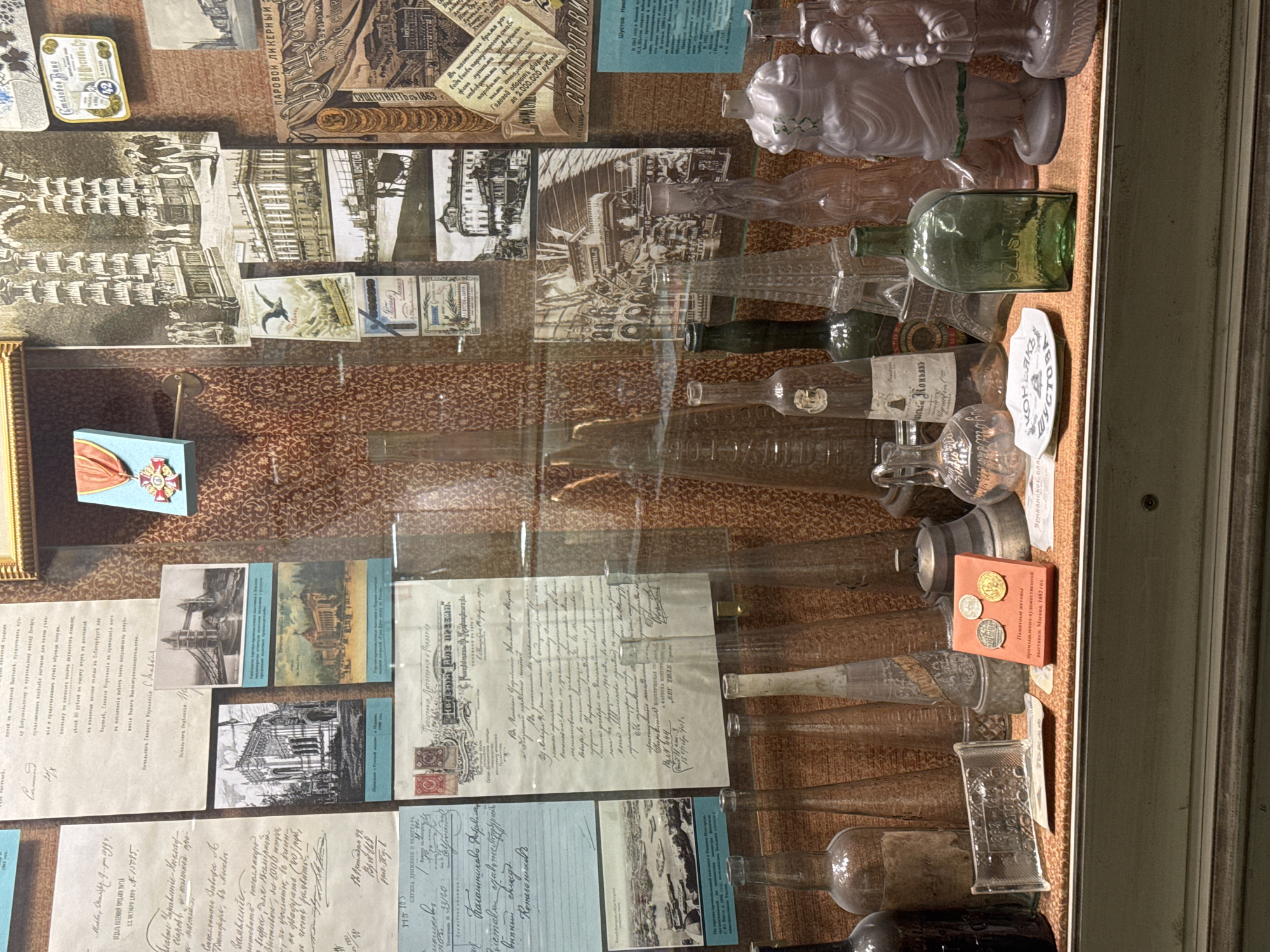 The image shows a display case containing a collection of historical documents, glassware, and other artifacts. The documents consist of handwritten letters and printed papers, possibly related to business or personal correspondence. Some of the documents feature stamps and handwritten signatures. The glassware includes bottles of various shapes and sizes, some of which are labeled. There is also a box of coins and medals commemorating an industrial-artistic exhibition in Moscow in 1882. The display case seems to be in a museum or historical archive, with a patterned background. The image is not related to Saint Petersburg, Russia.