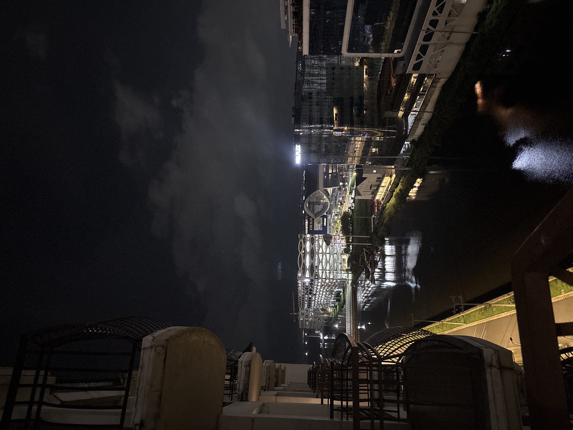 A nighttime view from a balcony shows a cityscape in Pasay, Philippines. A dark, reflective body of water, likely a canal or river, runs through the scene. Buildings with illuminated windows and striking architectural designs line the waterfront. One building features a unique, honeycomb-like facade, while another displays the "FLEXPLEX" logo illuminated in white. Construction cranes are visible in the distance, indicating ongoing development. The sky is dark and cloudy, with some lights from the city reflecting on the water's surface. The foreground shows parts of a balcony with railings and a worn concrete railing.
