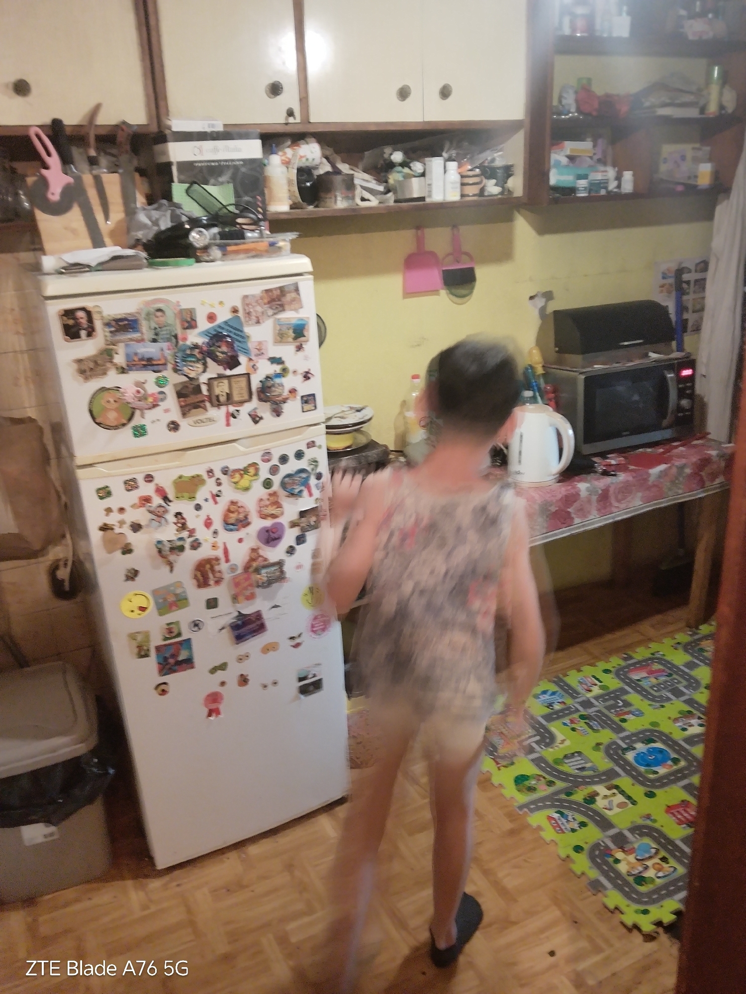 The image depicts the interior of a domestic kitchen or dining area in Stara Zagora, Bulgaria. A heavily motion-blurred individual, appearing to be a child or small adult, is present in the foreground, moving from left to right. The person is wearing a patterned sleeveless top, light-colored shorts or undergarments, and a dark slipper on the visible foot.
The environment includes light-colored wall cabinets with dark knobs positioned at the top of the left and center frames. Below the left cabinets, a wooden shelf holds kitchen implements such as a pink pizza cutter, knives in a block, and various boxes and bottles. A large white two-door refrigerator, heavily adorned with numerous magnets and stickers, occupies the left-center of the frame. To the immediate left of the refrigerator, a light gray trash can with a black liner is partially visible.
The wall behind the person is painted yellow. To the right, an open wooden shelving unit displays an assortment of bottles, boxes, and miscellaneous items, indicating a cluttered storage arrangement. A rectangular table, draped with a floral tablecloth featuring red/pink rose patterns on a light background, is positioned to the right of the person. On this table, a white electric kettle and a black microwave oven with illuminated red display numbers are visible. The floor is covered with light-toned wooden parquet flooring, transitioning to a green interlocking foam playmat with a road/cityscape design in the bottom right corner.