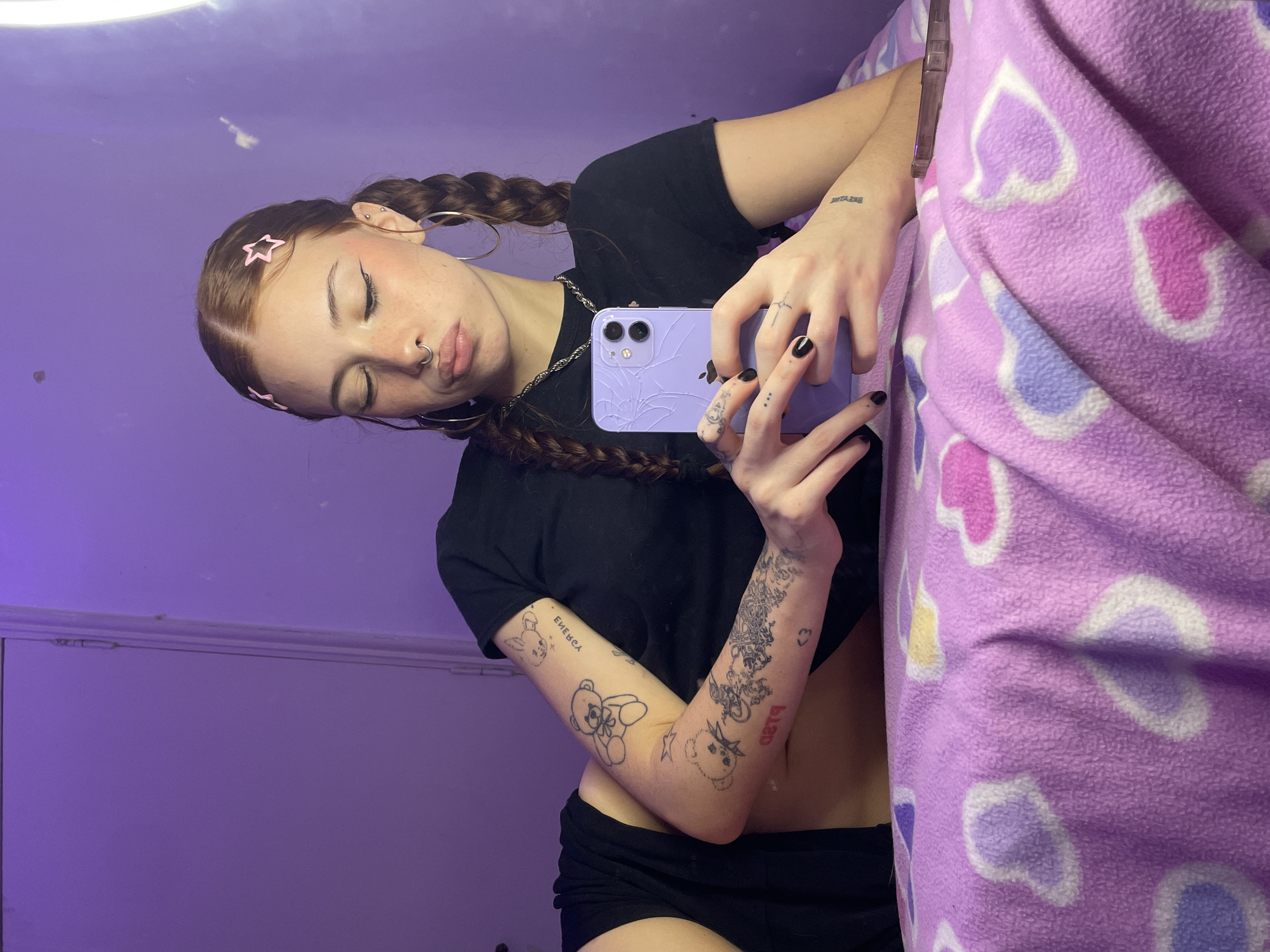 A young woman is taking a selfie in a room with a purple wall and a purple blanket with heart patterns. She has auburn hair styled in braids with pink star clips. She is wearing a black t-shirt and black shorts. She has multiple tattoos on her arms and hands. She is holding a purple smartphone with a cracked screen. She has a nose ring and large hoop earrings. A silver chain necklace is visible around her neck. The location appears to be indoors, possibly in a bedroom. There are no elements suggesting the image was taken in Buenos Aires, Argentina.