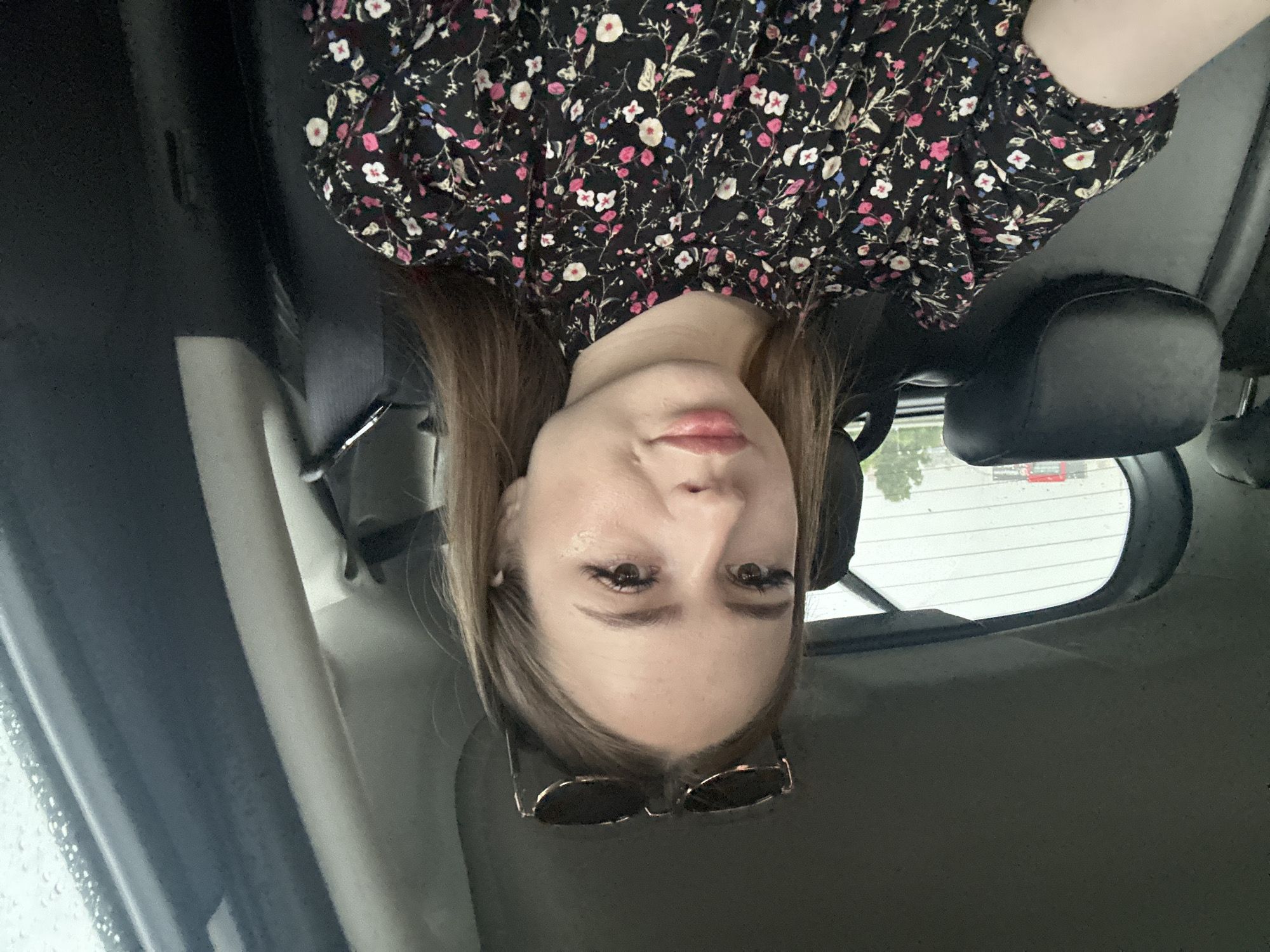 A young woman with blonde hair and sunglasses perched on her head sits in the passenger seat of a car. She is wearing a black floral print dress. The interior of the car is visible, including the seats and the ceiling. Outside the car, it appears to be daytime, and the background is blurred, suggesting movement. The overall scene is casual and appears to be a selfie taken while in transit.