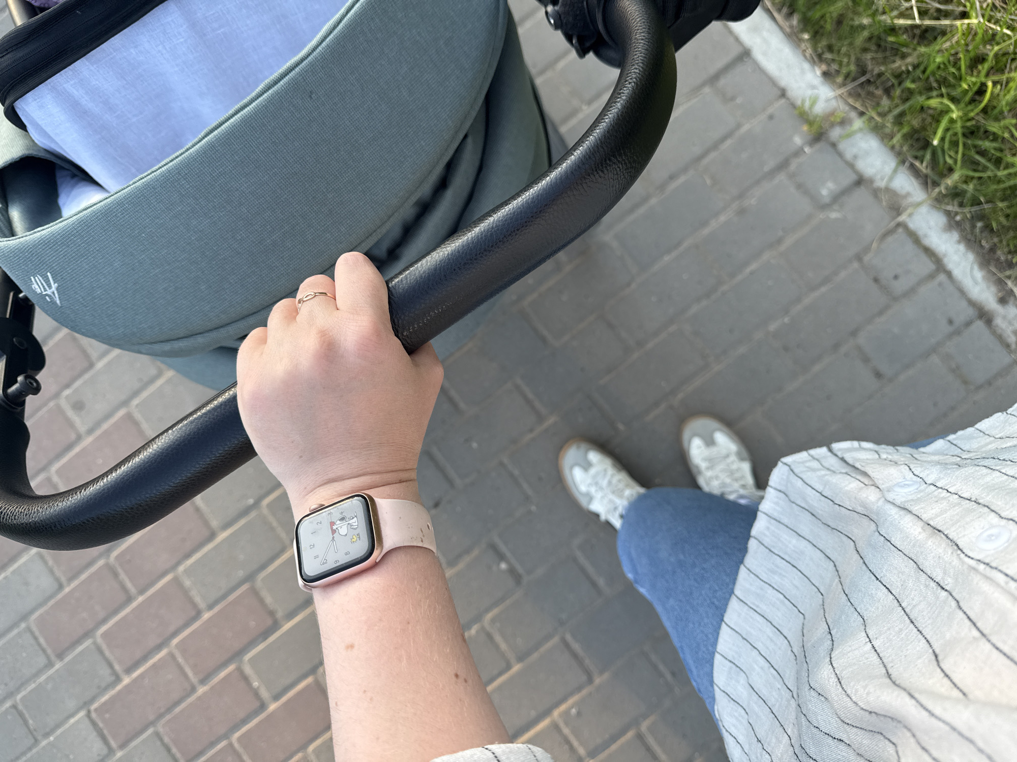 A first-person perspective shot shows a hand gripping the handle of a stroller. The hand has a light pink smartwatch on the wrist, displaying a Snoopy watch face. A thin gold ring is on the index finger. The person is wearing blue jeans and a white and black striped shirt. They are wearing white sneakers. The stroller has a sage green fabric. The scene is outdoors, on a paved walkway with a grassy edge. The location is unknown, likely in a city.