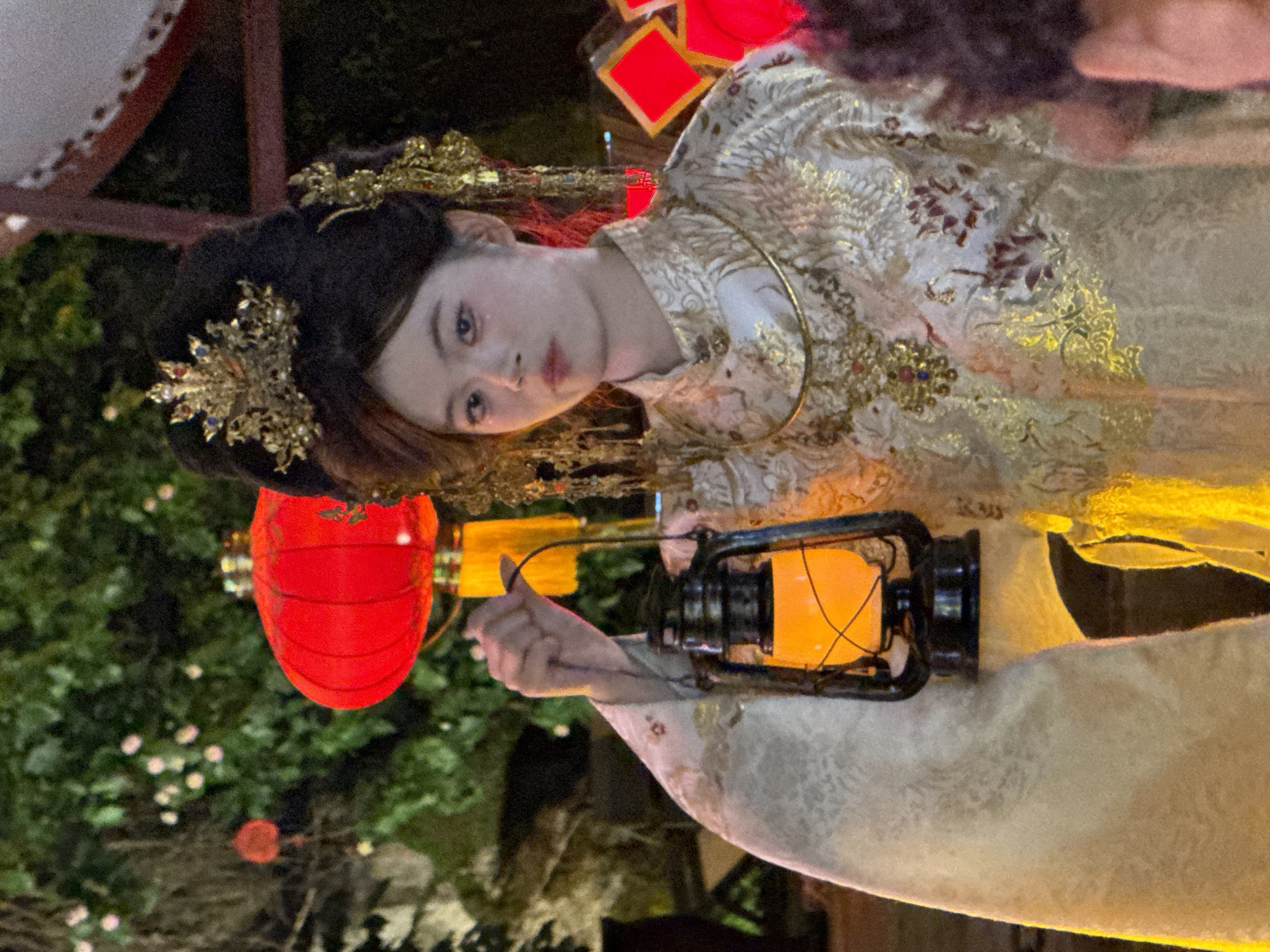 A young woman, with a traditional Chinese hairstyle adorned with gold ornaments and jewels, is holding a lit oil lantern in her left hand. She is wearing an elaborate, ornate gold and white brocade robe with floral patterns. Her makeup is light, with a touch of blush on her cheeks and a subtle hint of red on her lips. The background is dimly lit, with several red lanterns visible, suggesting a festive or cultural event. The scene appears to be outdoors at night, with foliage and what looks like a rocky outcrop in the background. The specific location in Yuzhong District, China, is not discernible from the image itself.