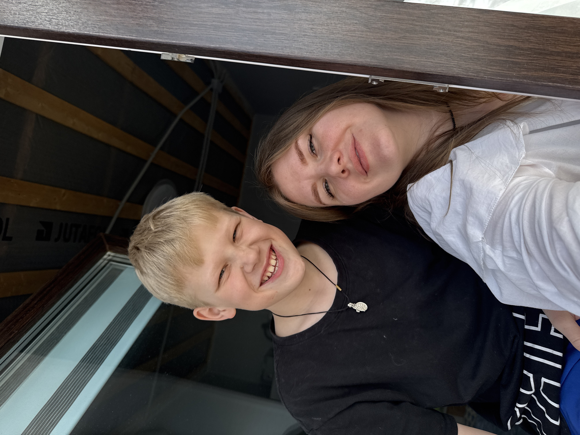 A close-up, eye-level photograph depicts two individuals, a blonde-haired male child and a brown-haired female adult, positioned within an indoor setting in Gornyi (Dvurechenskoe S/P), Russia. The male child, located on the left, exhibits a wide, toothy smile with crinkled eyes; he wears a black short-sleeved t-shirt with white graphic details visible on the lower portion, and a black cord necklace with a silver-colored oval pendant. The female, positioned to the right, displays a subtle smile and wears a white collared shirt and a dark choker necklace. A dark brown vertical frame, consistent with a window or door jamb, occupies the right edge of the image and includes a metallic latch mechanism near the female's shoulder. To the upper left, a window with partially open horizontal blue-grey blinds is visible. The ceiling above features dark panels interspersed with lighter, horizontal wooden or striped elements. Both subjects face the camera directly.