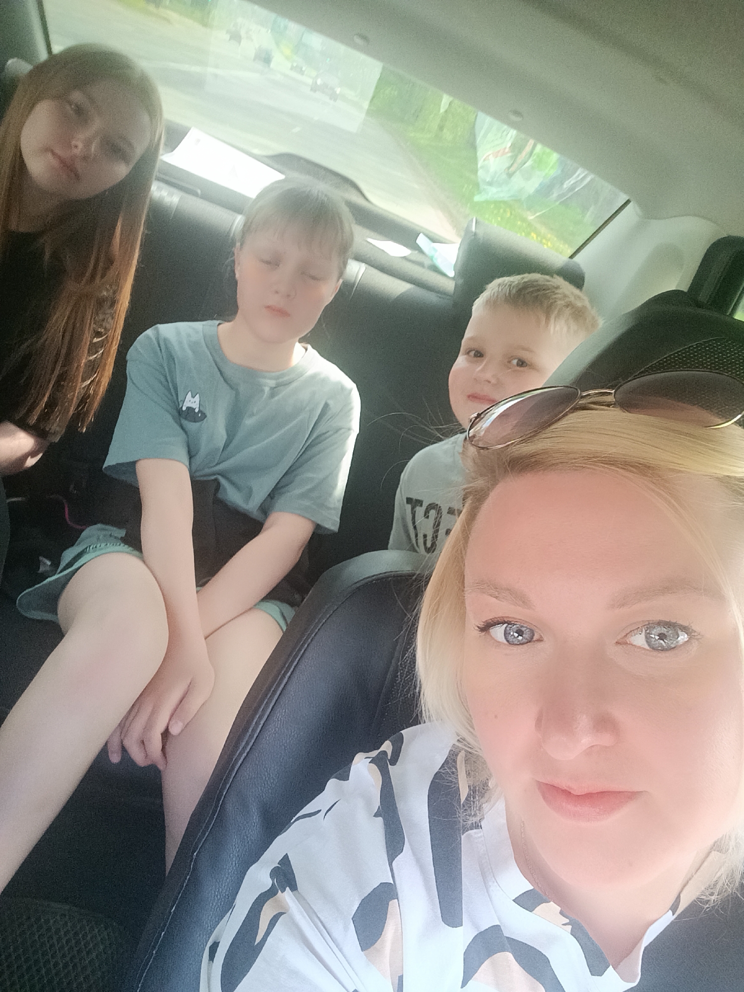 The image shows a selfie taken inside a car, likely a ride. Four individuals are visible: a woman with blonde hair and sunglasses perched on her head, presumably the one taking the photo, and three children in the back seat. From left to right in the back seat, there is a girl with long brown hair, a child with short blonde hair and eyes closed, and a young boy with blonde hair. The woman is wearing a patterned white shirt. The child in the middle is wearing a teal shirt with a cat design. The environment is the interior of a car, with glimpses of outside through the window, suggesting they are traveling. The exact location within the city of Chaikovsky, Russia is not identifiable from the visual information provided.