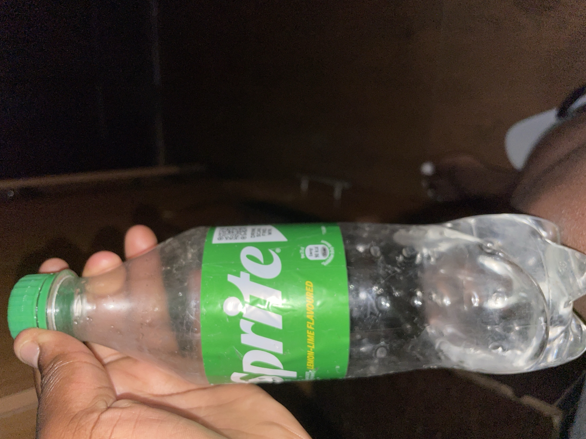 A person is holding a plastic bottle of Sprite in their hand. The bottle is mostly empty and has a green cap and a green label. The background is dark and indistinct. There is no specific information to indicate this image was taken in Kurunduma, Nigeria.