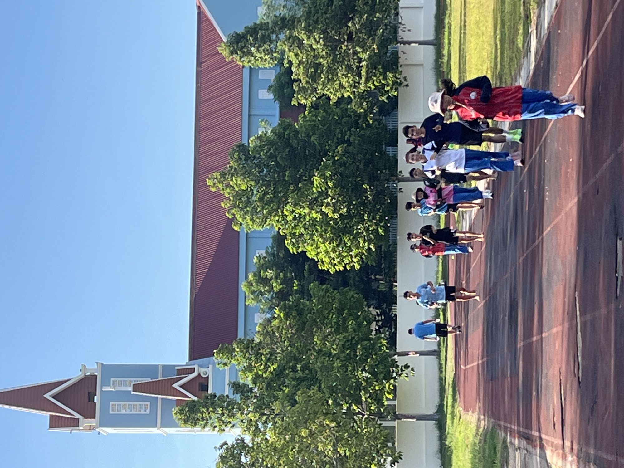In Phan Thiet, Vietnam, a group of individuals are running on a red track, possibly a sports field, in front of a building with a red roof and blue walls. The building features a tall tower with a red pointed roof. Green trees line the background, and the sky is clear blue. The runners are dressed in athletic wear, with some wearing casual clothing. One runner in the foreground is wearing a red jacket and blue pants. The track shows signs of wear and tear.