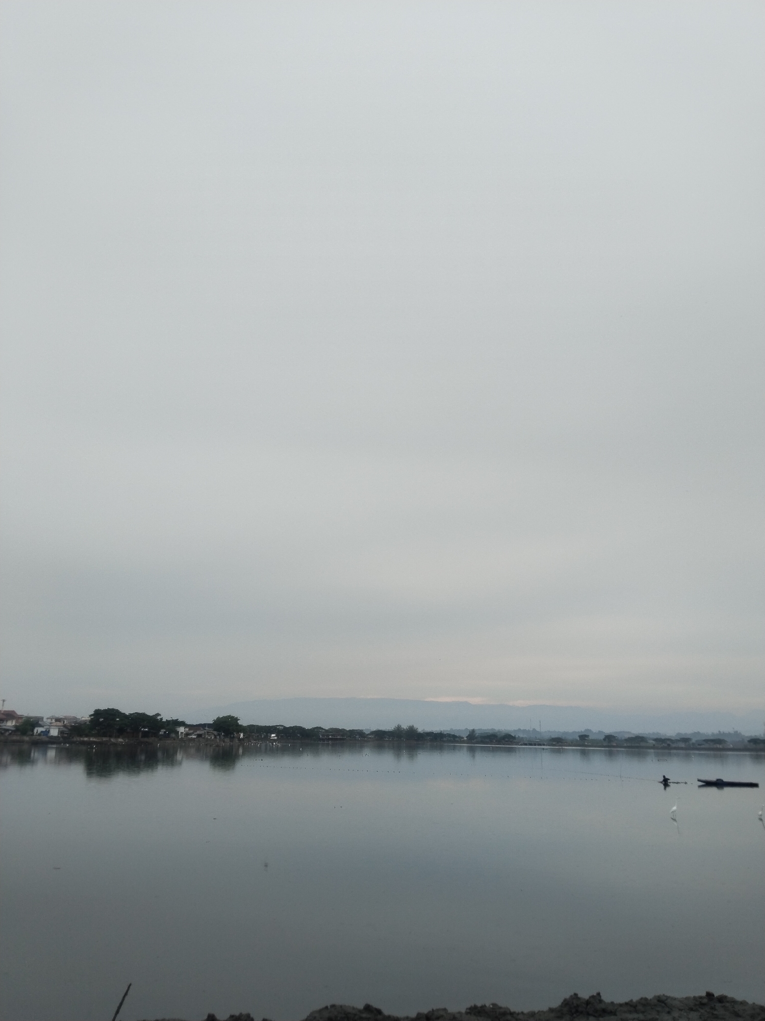 The image shows a serene view of the Lhokseumawe city lake at dawn. The sky is overcast, casting a soft, diffused light over the scene. The water is calm and reflects the sky and the distant shoreline. Along the shore, there are several houses and trees, and a line of poles stretches across the water. In the middle of the lake, a person is in a small boat, and two white birds are visible near the boat. The foreground shows a rocky embankment, with a few dry branches. The overall impression is one of tranquility and the beginning of a new day.