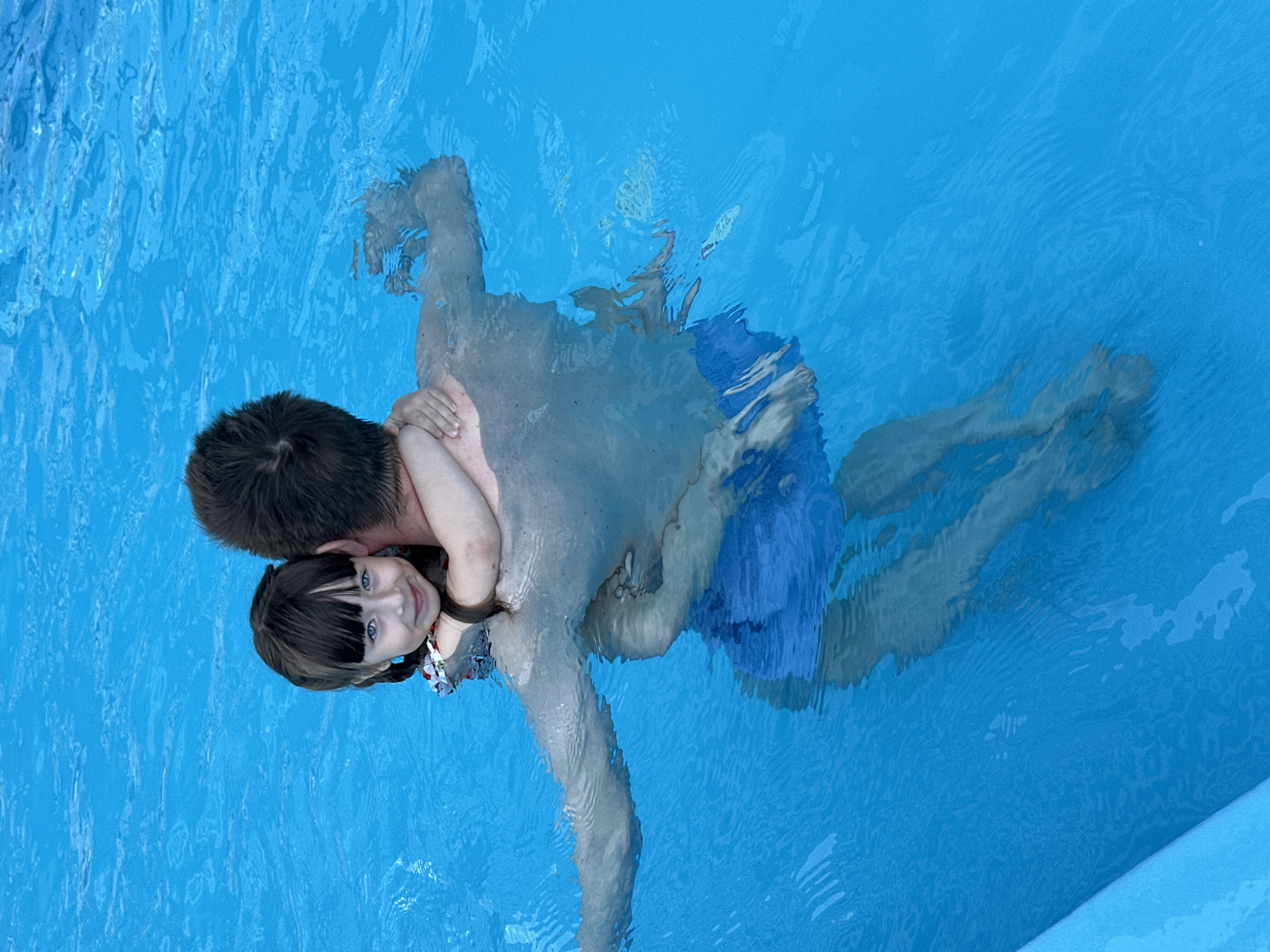 A top-down shot captures a man and a girl in a blue swimming pool. The girl, looking at the camera with a smile, has dark hair and blue eyes. The man, whose back is visible, is wearing blue swimming trunks. The pool water is clear, and the photograph seems to have been taken outdoors during daylight hours. The location is unknown; Sochi, Russia cannot be verified.