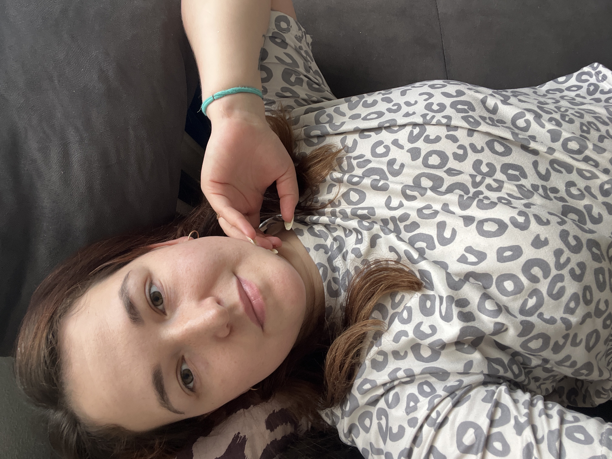 A close-up, high-angle shot features a young woman lying on a dark gray sofa. She is wearing a light-colored pajama set with a gray leopard print. Her head is tilted back and to her left, with her gaze directed slightly upwards towards the camera. Her right hand is resting on her chest, near her collarbone, and she has a thin, turquoise bracelet on her wrist. Her hair is brown and falls around her face and shoulders. The lighting appears to be natural, suggesting it could be daytime. The overall mood is relaxed and casual, implying the woman is at home in a comfortable setting in Chelyabinsk, Russia. No text is visible in the image.