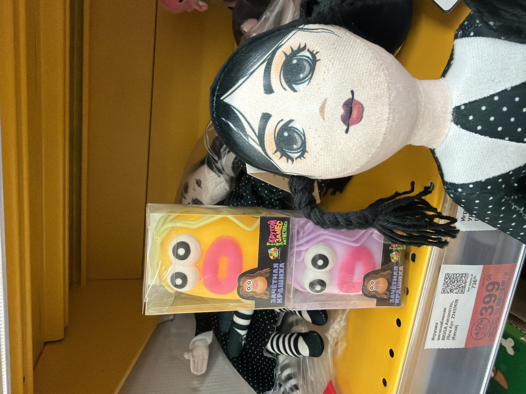 This media file presents a close-up view of merchandise within a retail setting, likely a toy aisle in a store located in Berezniki, Russia. The primary subject is a soft-stuffed doll, positioned horizontally, with a pale face, striking large eyes, small red lips, and black hair braided into pigtails. The doll is dressed in a black and white polka-dotted outfit, partially visible. Its overall aesthetic, characterized by its pale complexion and dark features, suggests a gothic or quirky style.
In front of the doll, two smaller, rectangular packaged items are displayed. These appear to be stress toys or squishies, one in yellow and the other in purple, each featuring large cartoon eyes and a pink "0" or "O" design. The packaging displays Russian text, including "Крутой ЗАМЕС АНТИСТРЕСС" (Cool MIX ANTISTRESS) and "ЗАЧЁТНАЯ КРАШИХА" (Cool CRUSH/BEAUTY).
To the right of the items, a retail price tag is visible. It identifies one of the items (likely the large doll) as "Игрушка мягконабивная BIGGA Аксолотль, 28см" (Toy soft-stuffed BIGGA Axolotl, 28cm) from China. The price tag shows a discounted price of 399.00 Russian Rubles, marked down by 45% from an original price of 736 Rubles. The background consists of solid yellow shelving, typical of a store interior, illuminated by artificial lighting. No individuals are present, and the scene focuses entirely on the products on display.
