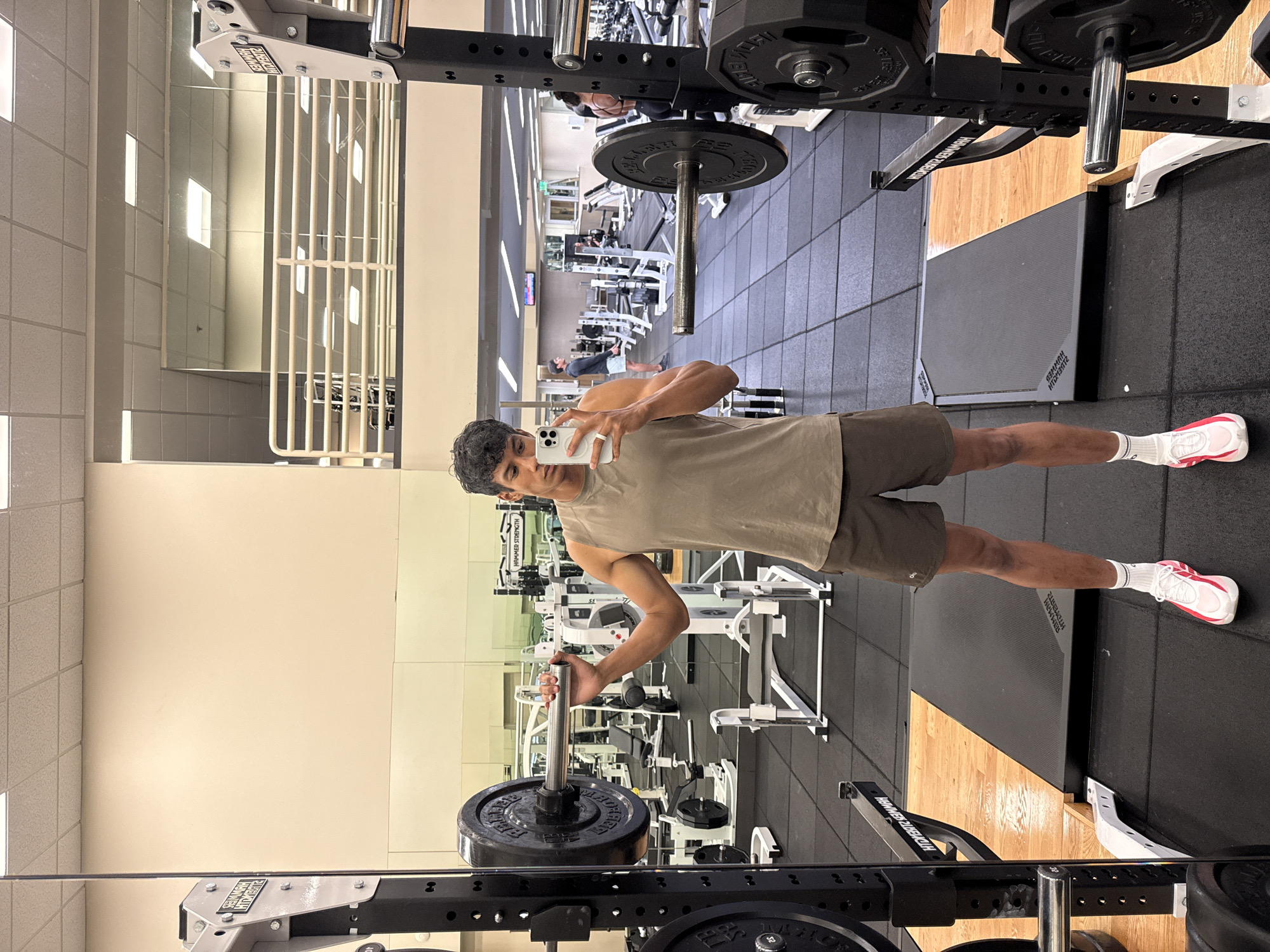 A man stands in a gym, holding a phone in his right hand and a barbell with weights in his left hand. He is wearing a light brown tank top, dark brown shorts, white socks, and white and pink athletic shoes. The gym has various workout machines and weightlifting equipment, with mirrors along the walls. The floor is tiled with black rubber squares and interspersed with wooden planks. Fluorescent lights illuminate the gym. The image reflects in the mirrors and is likely a selfie taken by the man. It cannot be determined whether or not the location is in Los Angeles, United States of America.