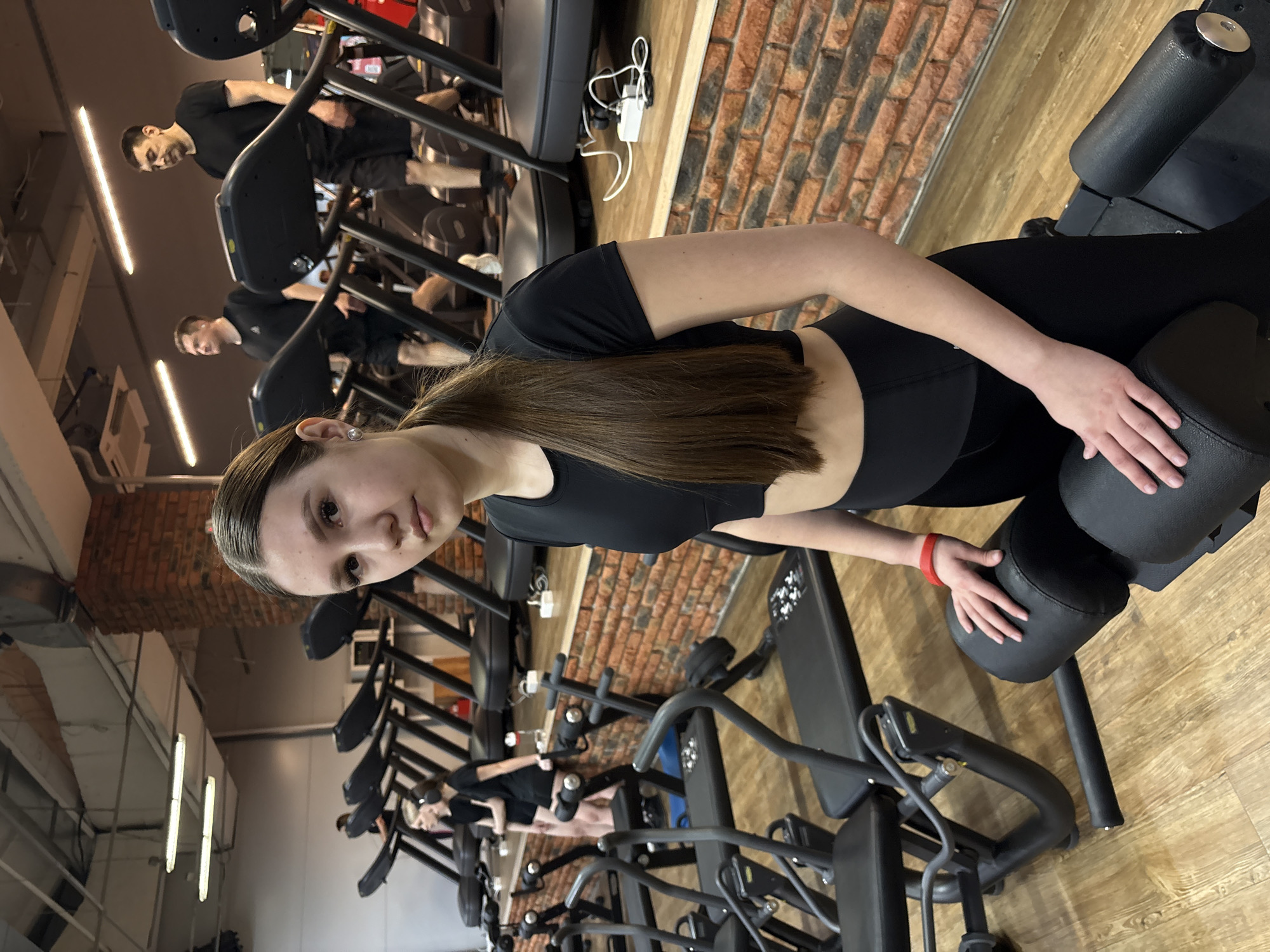 This image captures a young woman at a gym in Chaikovsky, Russia, presented with a 90-degree clockwise rotation. The main subject, a light-skinned woman with long brown hair, is looking directly at the camera with a neutral expression. She is wearing a black short-sleeved crop top and black leggings, and has a red wristband on her left wrist. She is seated on a black gym machine, resting her hands on its padded surfaces. A small, circular earring is visible in her right ear.
In the background, several other individuals, mostly men, are engaged in exercise. They are seen on treadmills and stair-stepper machines, dressed in athletic wear. The gym features an industrial-style aesthetic with a prominent exposed red brick wall, light-colored wooden flooring, and light gray painted walls. Bright fluorescent strip lights illuminate the indoor setting. Various other pieces of gym equipment are scattered throughout the facility, including benches and weight machines. Power cables and an adapter are visible near one of the treadmills.
The scene depicts a typical gym environment during operational hours, likely daytime or evening, given the bright interior lighting. No specific activity is shown for the main subject other than resting or posing on the machine, while the background figures are actively working out. There is no discernible text visible on any equipment or surfaces within the frame. The overall atmosphere is focused on fitness and exercise.