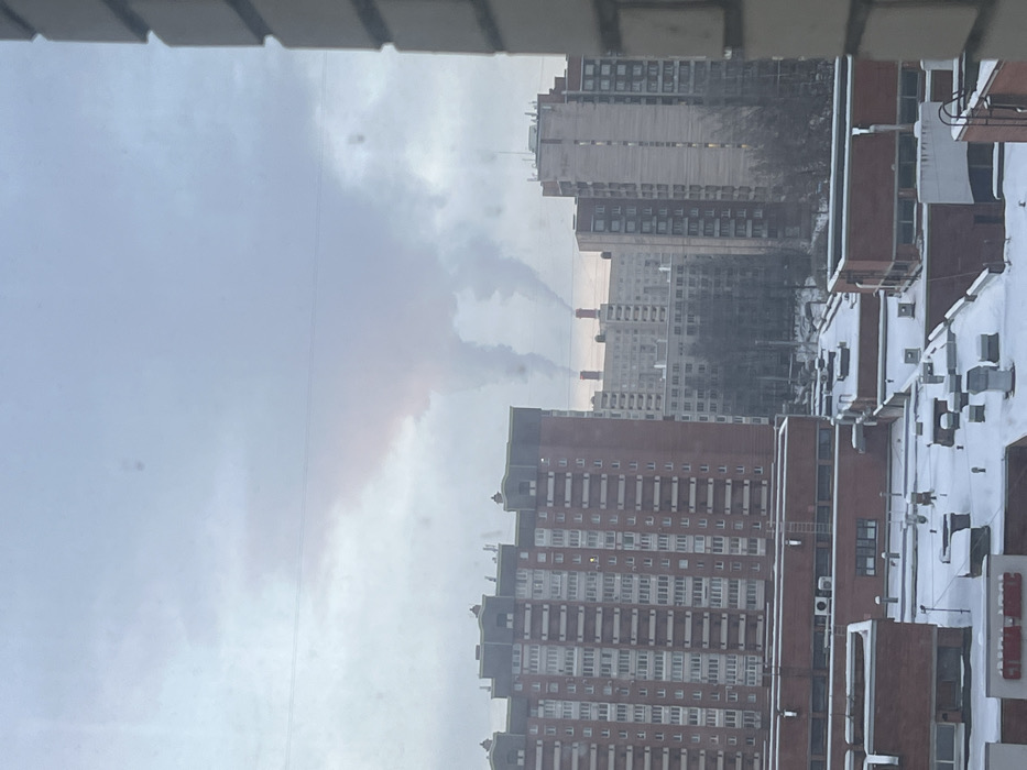 A view from a window shows a cityscape under a cloudy sky. Several tall apartment buildings with numerous windows dominate the scene. Some buildings appear older with a faded gray color, while others have a red brick facade. Smoke or steam rises from structures in the distance, perhaps chimneys or cooling towers. The foreground includes parts of the building from which the photo was taken, adding a sense of depth. The overall impression is urban, possibly in a post-industrial setting.