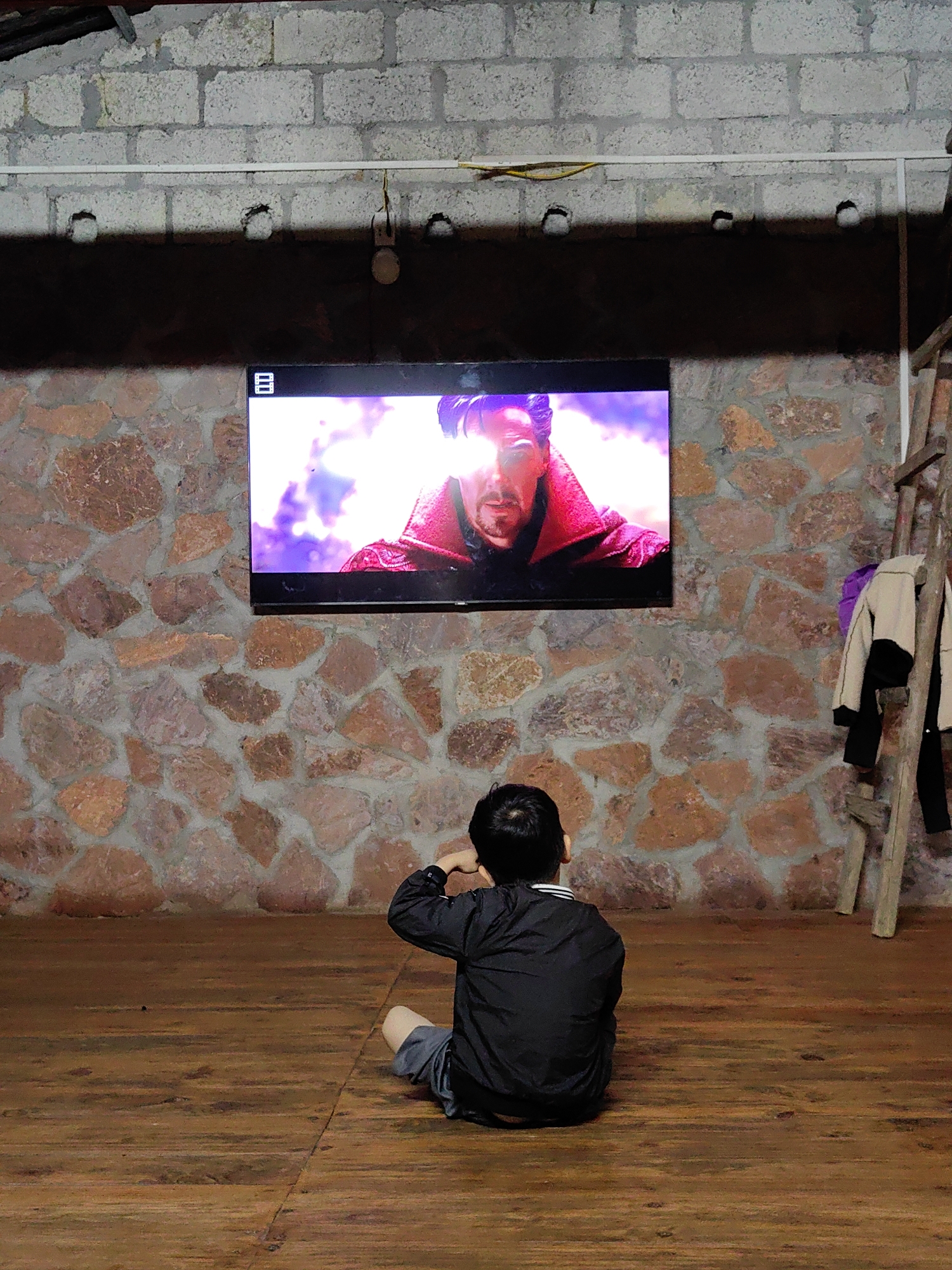 A boy sits on a wooden floor watching a television mounted on a stone wall. The TV displays a scene featuring a man with light emanating from his face. A wooden ladder leans against the wall to the right of the television, with clothes hanging on it. Above the TV, the wall transitions to concrete blocks. The photo was taken in Xã Đàm Thủy, Vietnam.