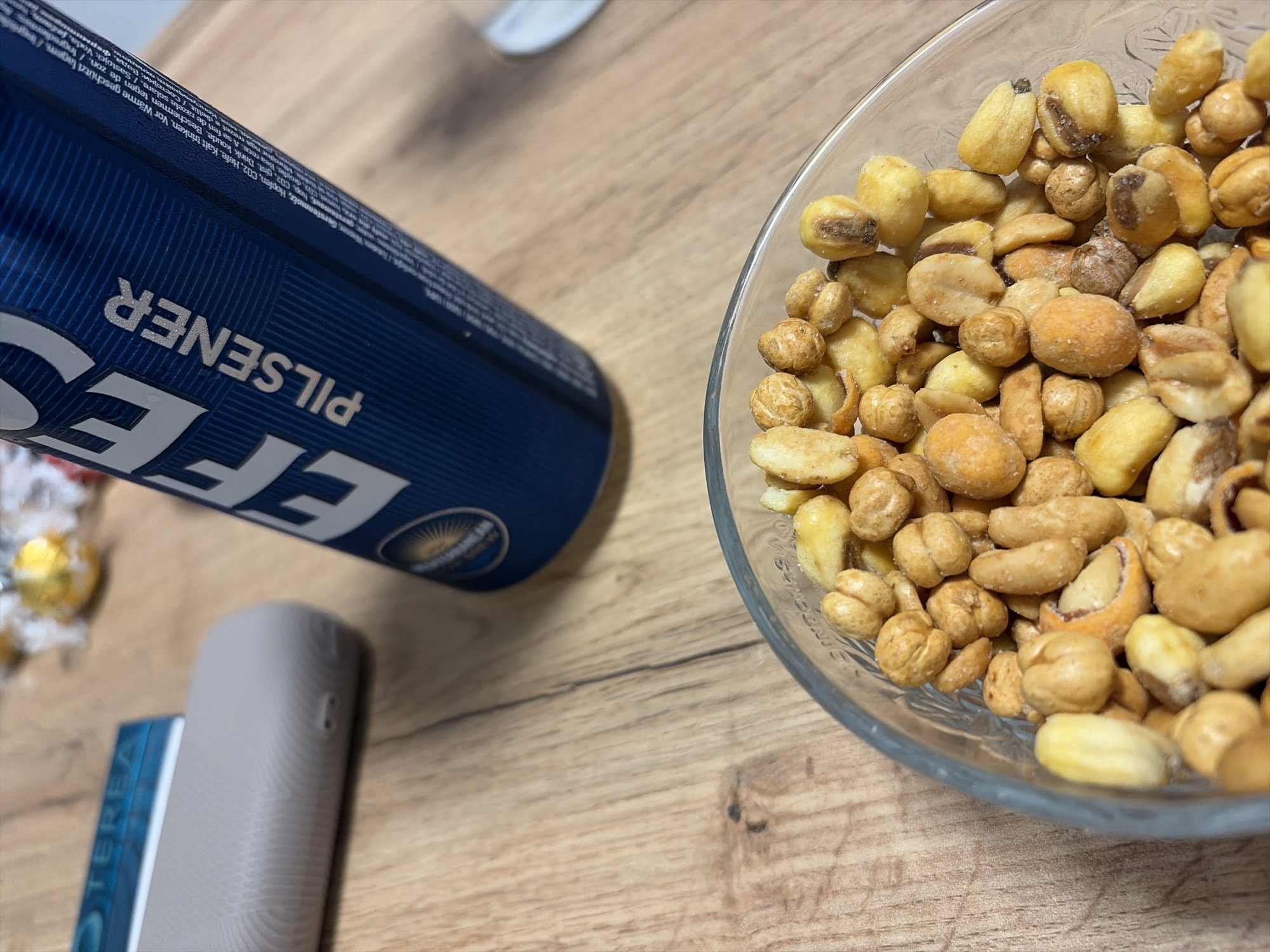 A high-angle shot captures a scene on a wooden table. A blue can of Efes Pilsener beer is positioned to the left, partially out of focus. Adjacent to the can is a gray, rectangular device, possibly a vape pen. A clear glass bowl filled with a variety of roasted nuts dominates the right side of the image. The lighting suggests an indoor setting. There is no visible indication of the image being taken in Dulovo, Bulgaria.