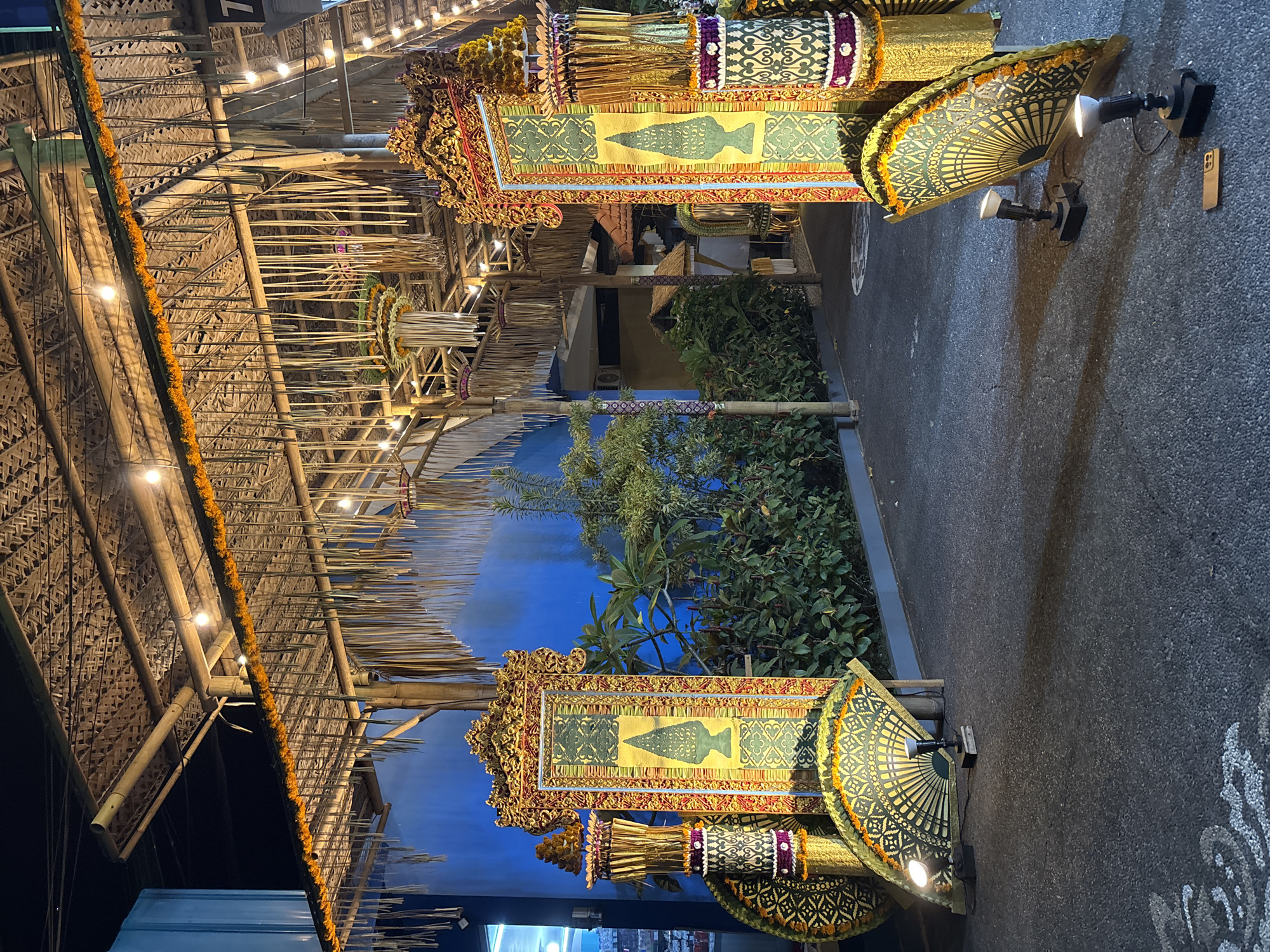 The image depicts an outdoor setting in Pecatu, Indonesia, at night. There is a structure overhead, constructed from bamboo and woven palm leaves, and decorated with strings of orange flowers and small lights. Along a paved walkway are two decorative displays, featuring framed artwork with ornate gold borders and images of what appears to be traditional vases. These displays also incorporate parasols, floral arrangements, and patterned fabrics. Behind the displays, there are bushes and a blue wall. There are also wall-mounted spotlights illuminating the scene.