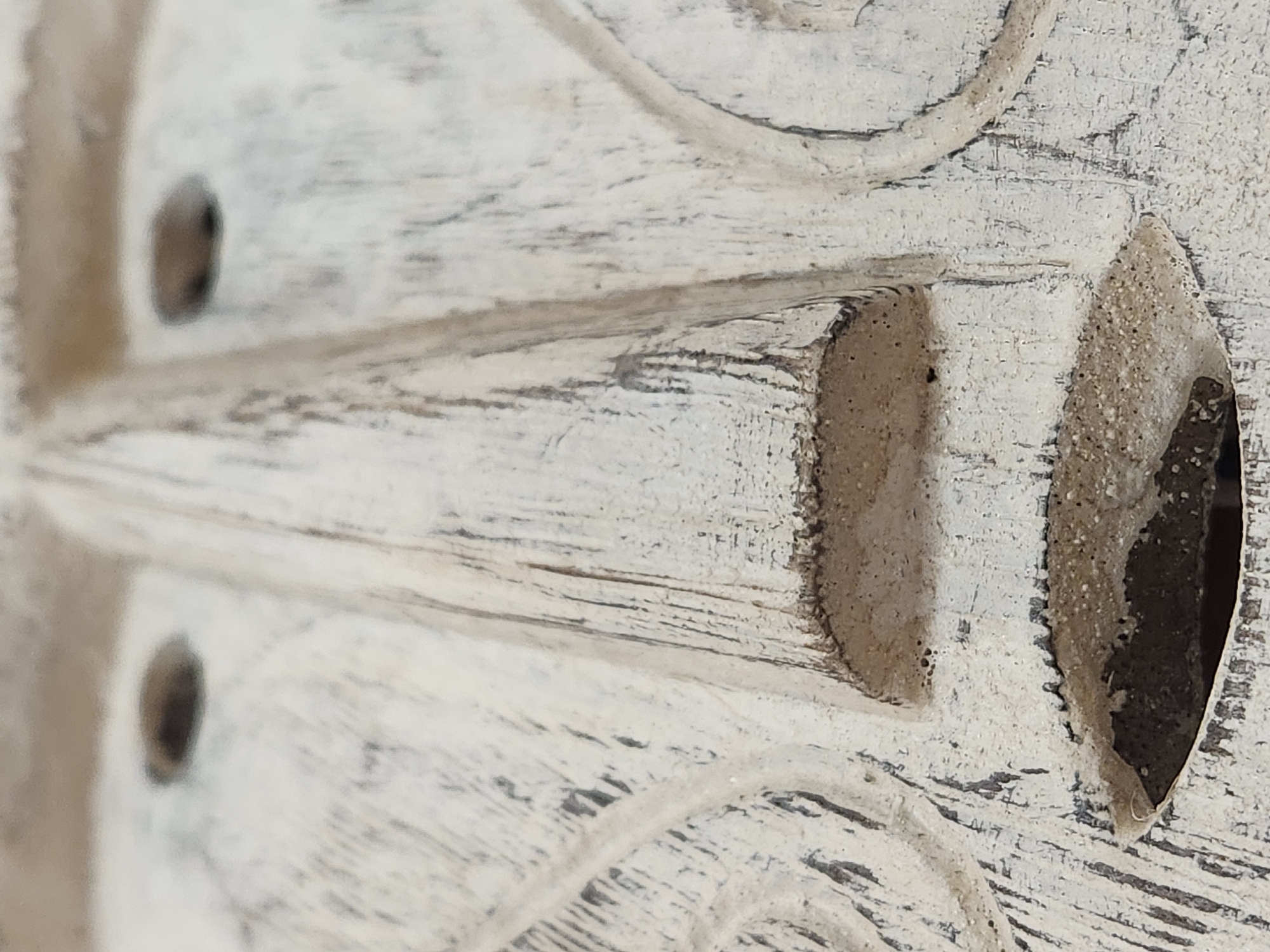 A close-up, shallow depth of field photograph shows a portion of a carved wooden mask, likely a decorative or cultural artifact. The mask is painted white with dark, aged undertones visible through the paint, suggesting wear and texture. The central focus is on the nose and mouth area of the mask. The nose is prominent and elongated, with a slight curve. The mouth is open, revealing a dark interior. The surface of the mask exhibits visible wood grain and fine cracks, contributing to its rustic appearance. The lighting is diffused, creating soft shadows and highlighting the texture of the wood. The overall impression is one of an aged, handcrafted object. There is no identifiable city, state, country, or specific location present in the image or derivable from the visual information.