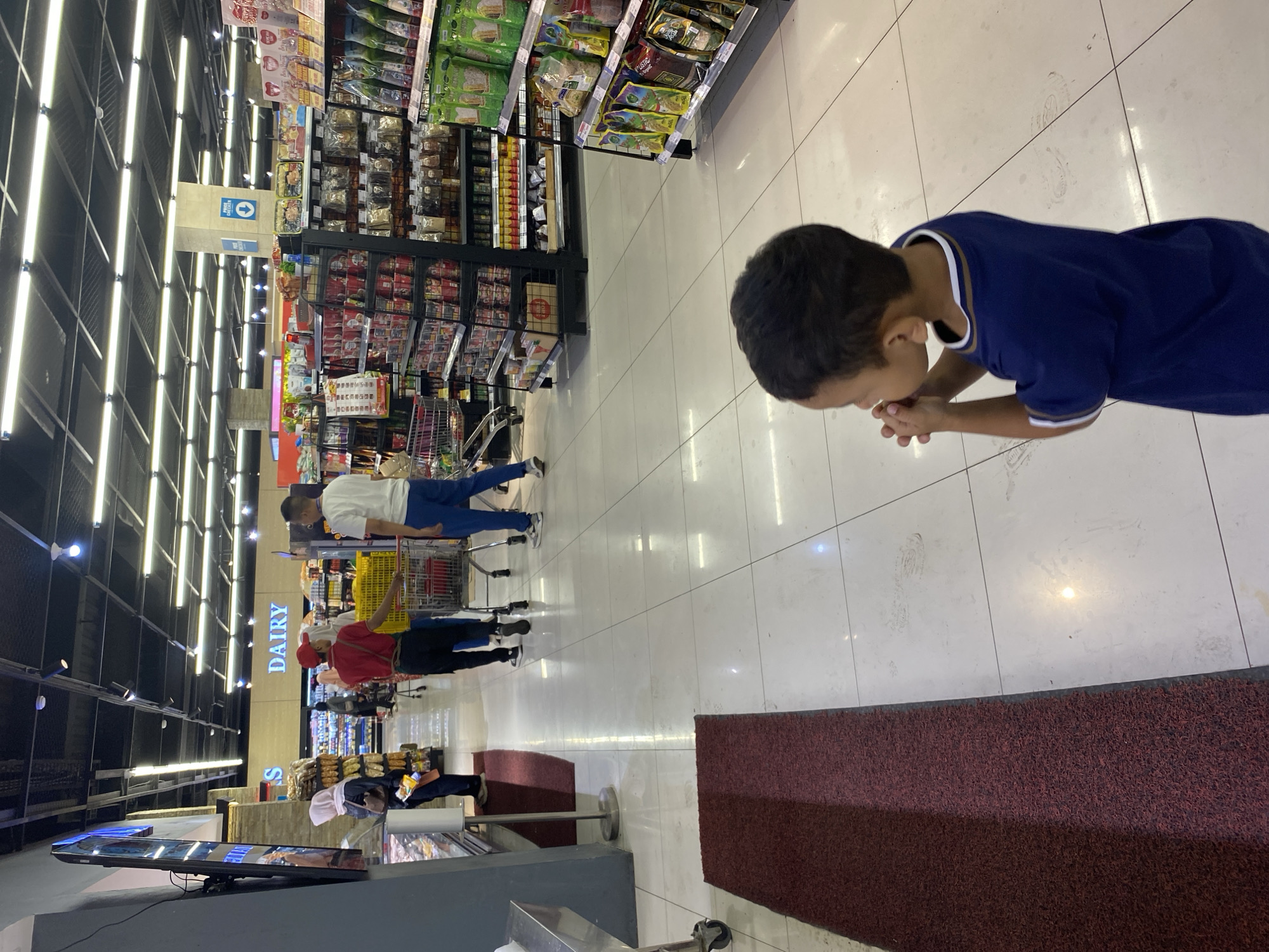 The image depicts the interior of a well-lit supermarket in Banda Aceh, Indonesia. In the foreground, a young boy in a blue t-shirt stands with his hands clasped, appearing to be looking at something off-camera. In the mid-ground, several shoppers are visible. A woman in a pink hijab and a man in a red cap are pushing a shopping cart towards the right. Another man in a white t-shirt and blue pants is walking towards the left. The supermarket aisles are stocked with various products, including what appears to be dairy items, packaged foods, and beverages. The floor is tiled, and there are overhead fluorescent lights illuminating the space. Red floor mats are placed in strategic locations.