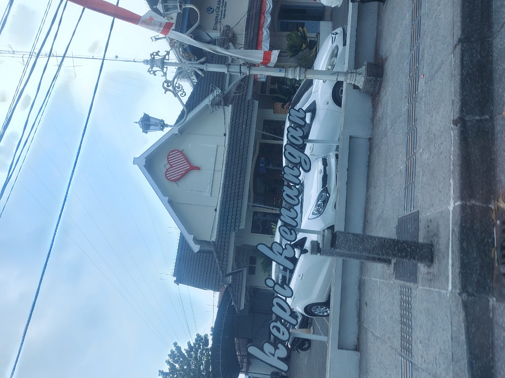 The image depicts the exterior of a building in Yogyakarta, Indonesia, with a prominent sign that reads "kopi kenangan." The building itself is light-colored with a dark tiled roof and features a decorative heart-shaped logo on its facade. In front of the building, two white cars are parked. A street lamp with decorative metalwork stands to the right of the building, and a red and white flag is visible, also to the right. The foreground shows a paved sidewalk with tactile paving for the visually impaired, indicating a public area within the city. The sky is overcast.