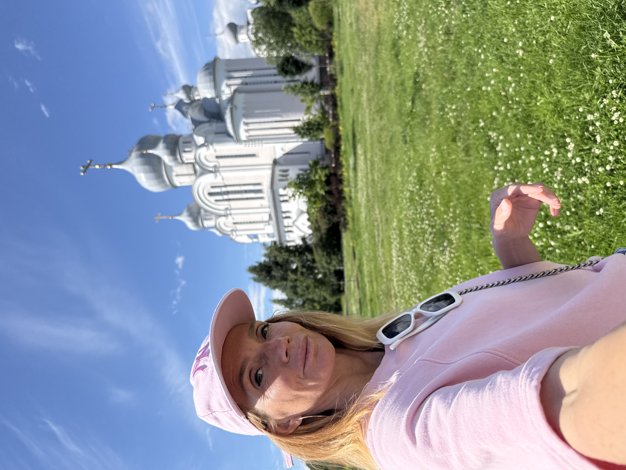 In Saint Petersburg, Russia, a blonde woman wearing a pink baseball cap and a pink sweatshirt takes a selfie. She is outdoors, with a grassy field dotted with small white flowers in the foreground. In the background, a large, white Orthodox church with multiple silver domes and crosses stands against a bright blue sky with wispy clouds. She has white sunglasses hanging from her sweatshirt. The woman is smiling slightly.