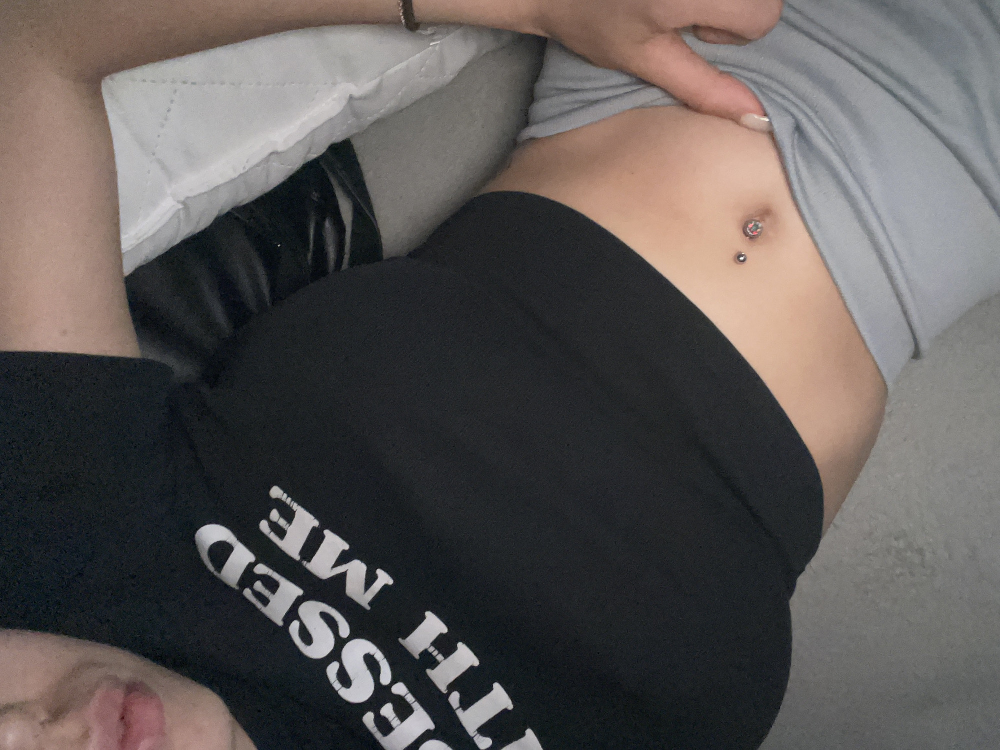 The image shows a close-up, overhead view of a person lying down. The person is wearing a black t-shirt with white text that reads "BLESSED WITH ME". They are also wearing black leggings and a grey top. The person has a belly button piercing with a sparkly barbell and a smaller stud below it. The background appears to be a bed with a grey pillow. The lighting is soft and appears to be indoor lighting. The focus is on the person's midsection and stomach. The overall mood is relaxed and casual. There are no obvious indicators of time of day, weather, or specific location beyond it being indoors.
