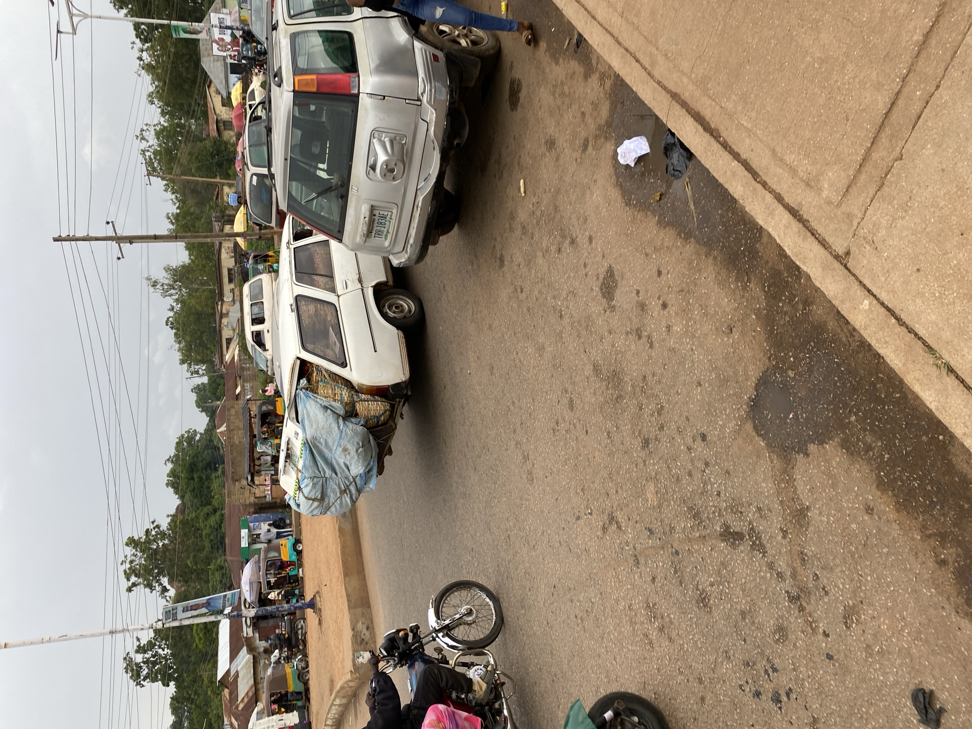 The scene is a street in Bukuru, Nigeria. A row of parked vehicles line the street, including a white van covered with a blue tarp and a silver SUV with a license plate that appears to read "TRR-783AE." People and vehicles are present further down the road. A motorcycle is parked on the side of the road in the foreground. Trash is visible near the curb. Overhead power lines are also present. The weather appears overcast.