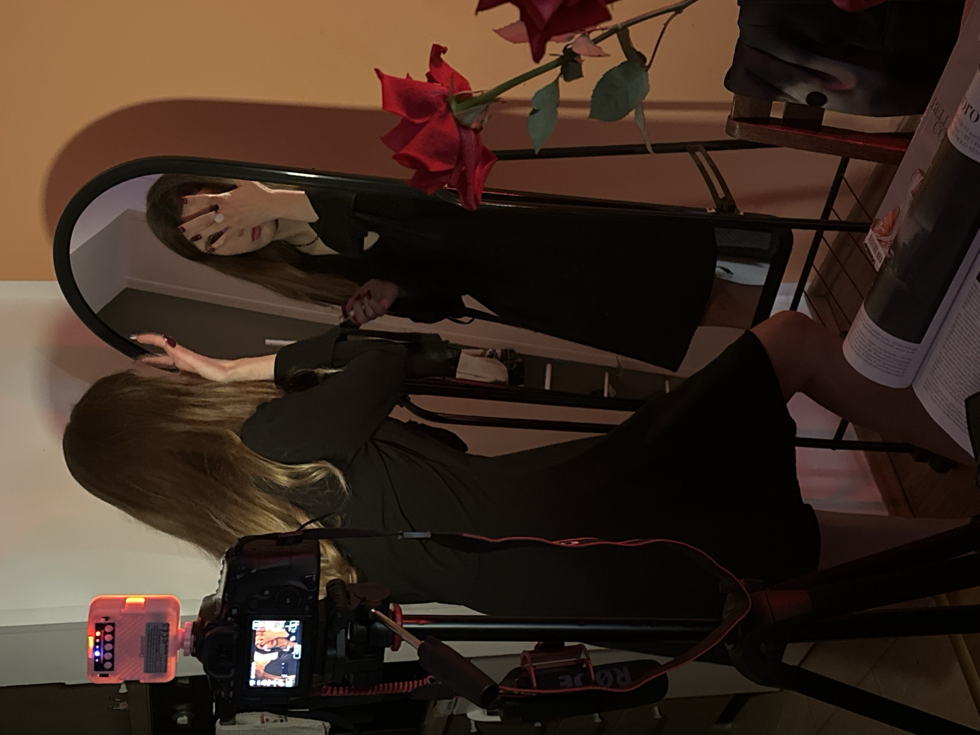 A woman in a black dress is being filmed by a camera. The camera is equipped with a microphone and a light. The woman is posing with one hand covering her face in a mirror, and the other hand outstretched towards the camera. There are red roses in the scene. A magazine rests on the chair next to the woman. The location cannot be determined, as the city of Šušanj, Montenegro is not recognizable in the scene.