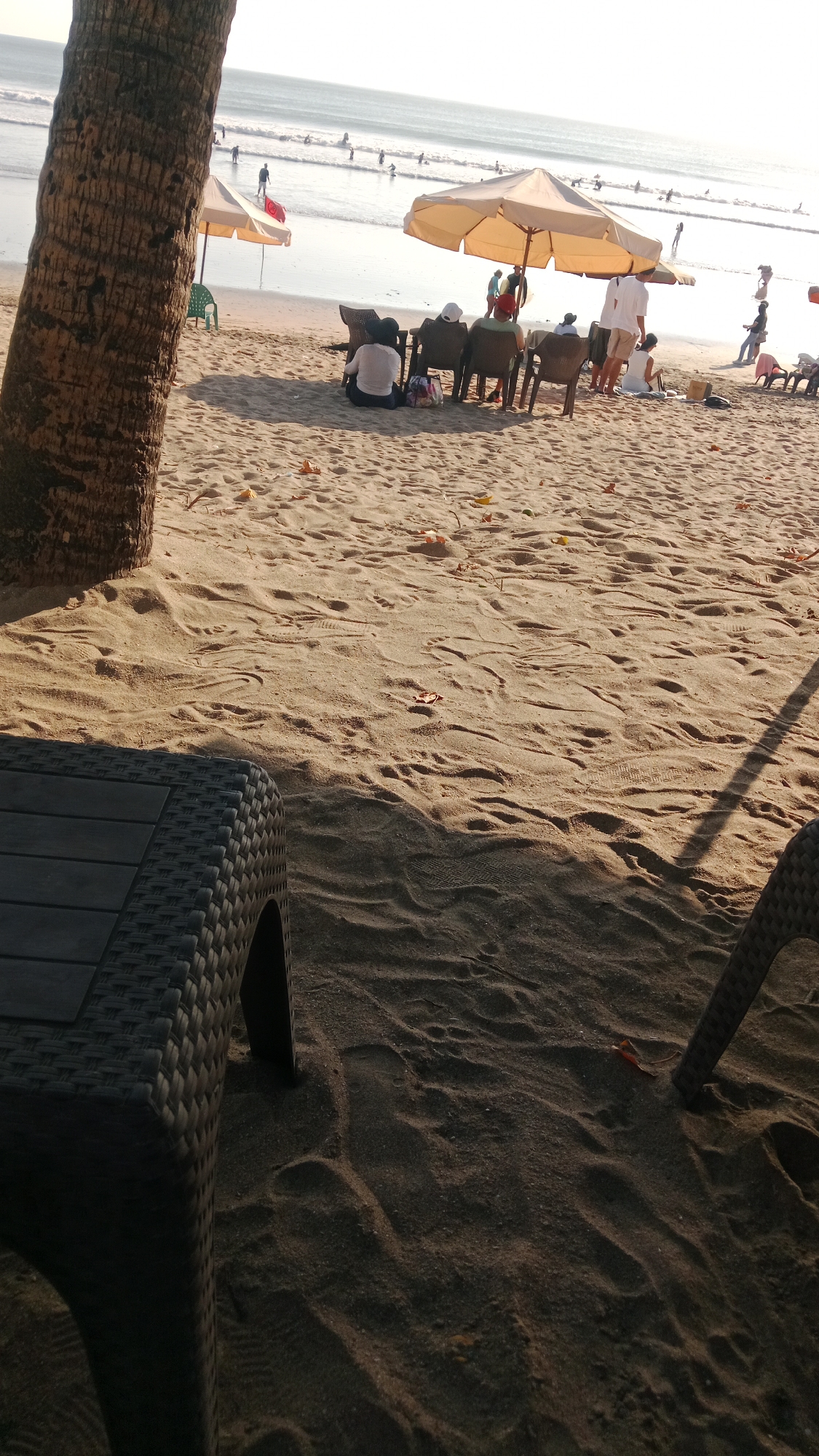 This image captures a beach scene in Kuta, Indonesia, likely during daylight hours given the lighting conditions. In the foreground, a portion of a dark-colored, woven-texture table or chair is visible, casting a shadow onto the sand. The sand itself is textured with footprints and scattered leaves or debris.
Beyond the foreground, several beach chairs are arranged on the sand, some occupied by people. One group appears to be sitting and facing the ocean. An umbrella provides shade over some of the chairs. Further back, the ocean stretches to the horizon, with gentle waves and numerous individuals visible in the water, some appearing to be swimming or surfing. Beach umbrellas and a few more people are scattered along the shoreline. A palm tree trunk is prominent on the left side of the image, partially obscuring the view. The overall impression is a relaxed beach atmosphere with various people enjoying the seaside environment.