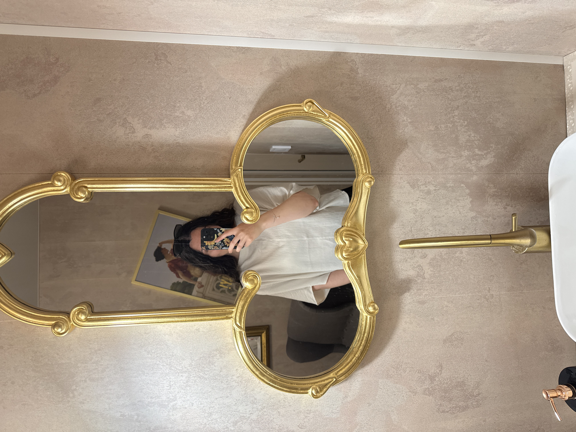 A person is reflected in a gold-colored mirror, which is mounted on a wall in a bathroom. The person is holding a smartphone, presumably taking the picture. The mirror also reflects a framed picture and other objects in the room. The wall behind the mirror appears to be textured and beige, with a white trim. A brass-colored faucet and a white sink are visible to the side of the mirror. The specific location and city are unknown.