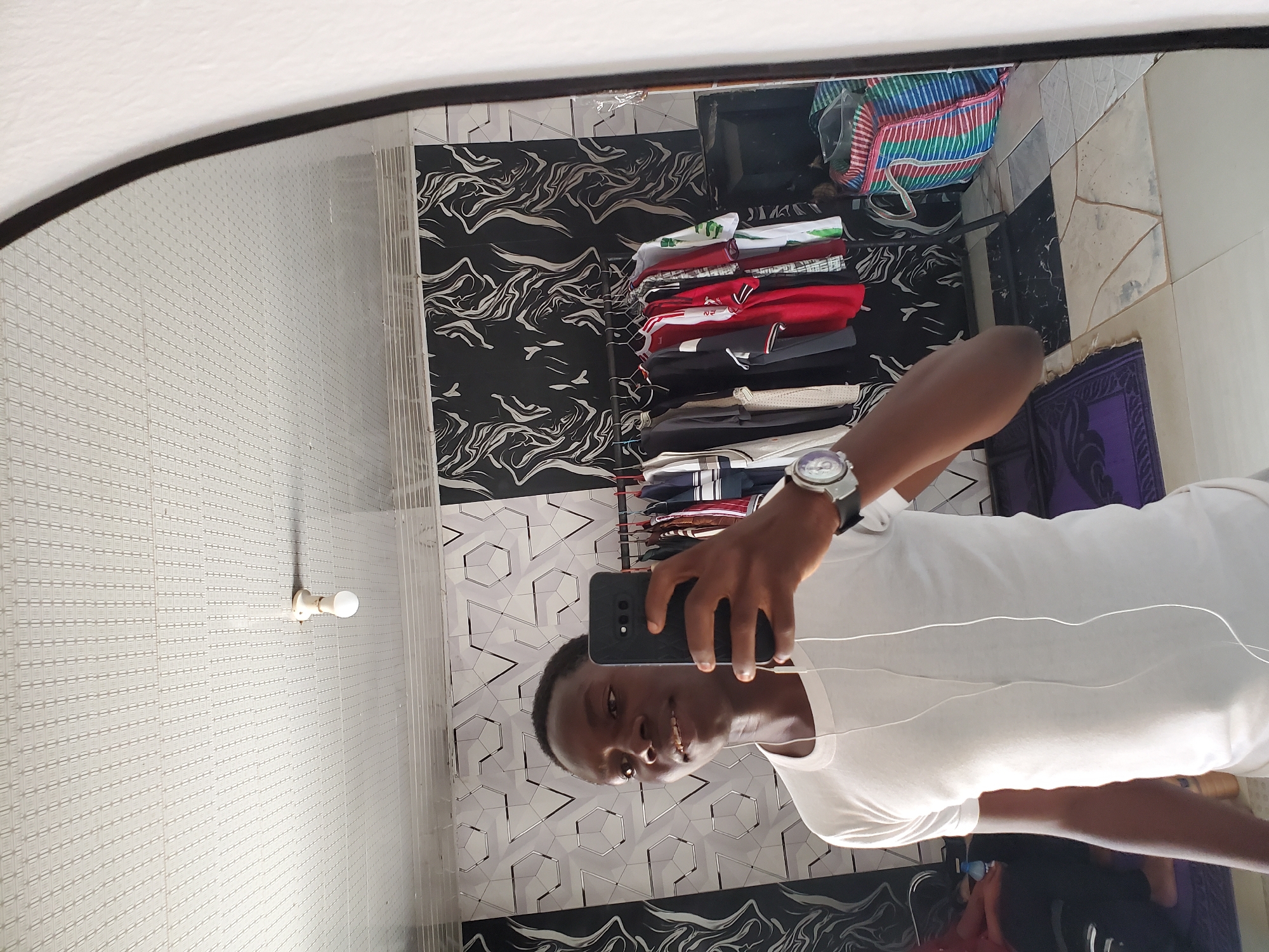 The image depicts a man taking a selfie in a mirror. He is wearing a white t-shirt and a watch. He is holding a black smartphone. The background includes patterned wallpaper in black and white, a rack of folded clothes, and a bag. The location is suggested to be Jos, Nigeria.