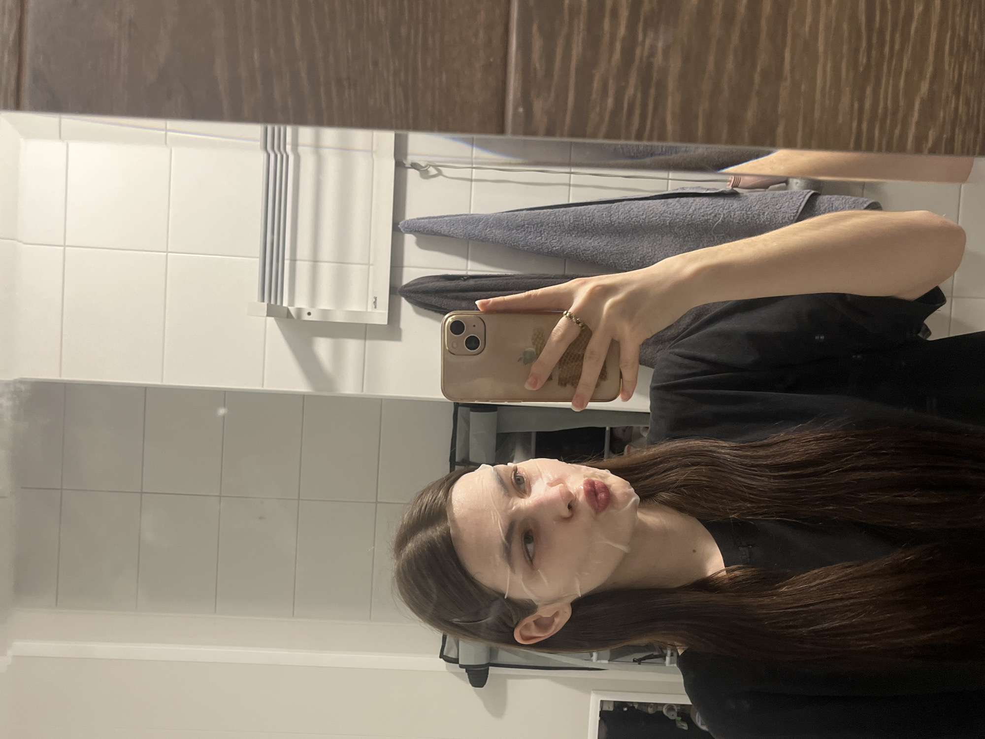 The image depicts a young female subject taking a selfie via a reflection in a bathroom mirror, purportedly located in Saint Petersburg, Russia. The subject has long, straight dark brown hair, parted centrally and secured with two white clips on either side of her head. She is wearing a white facial sheet mask covering her forehead, nose, cheeks, and chin. Her lips are pursed into a distinct "kissing" gesture. She is dressed in a dark, likely black, long-sleeved top. Her right hand, adorned with a ring on the ring finger, holds a gold-colored smartphone, featuring a dual-camera system, directed towards the mirror to capture the photograph. The reflected bathroom environment features white rectangular wall tiles. Mounted on the tiled wall is a white horizontal towel warmer with multiple bars. Below the warmer, two towels, one dark grey and one light grey/blue, hang from hooks. A dark, indistinct object is partially visible on the wall to the left of the subject's reflection. To the right, a section of wooden cabinetry or door frame with a vertical grain pattern is reflected.