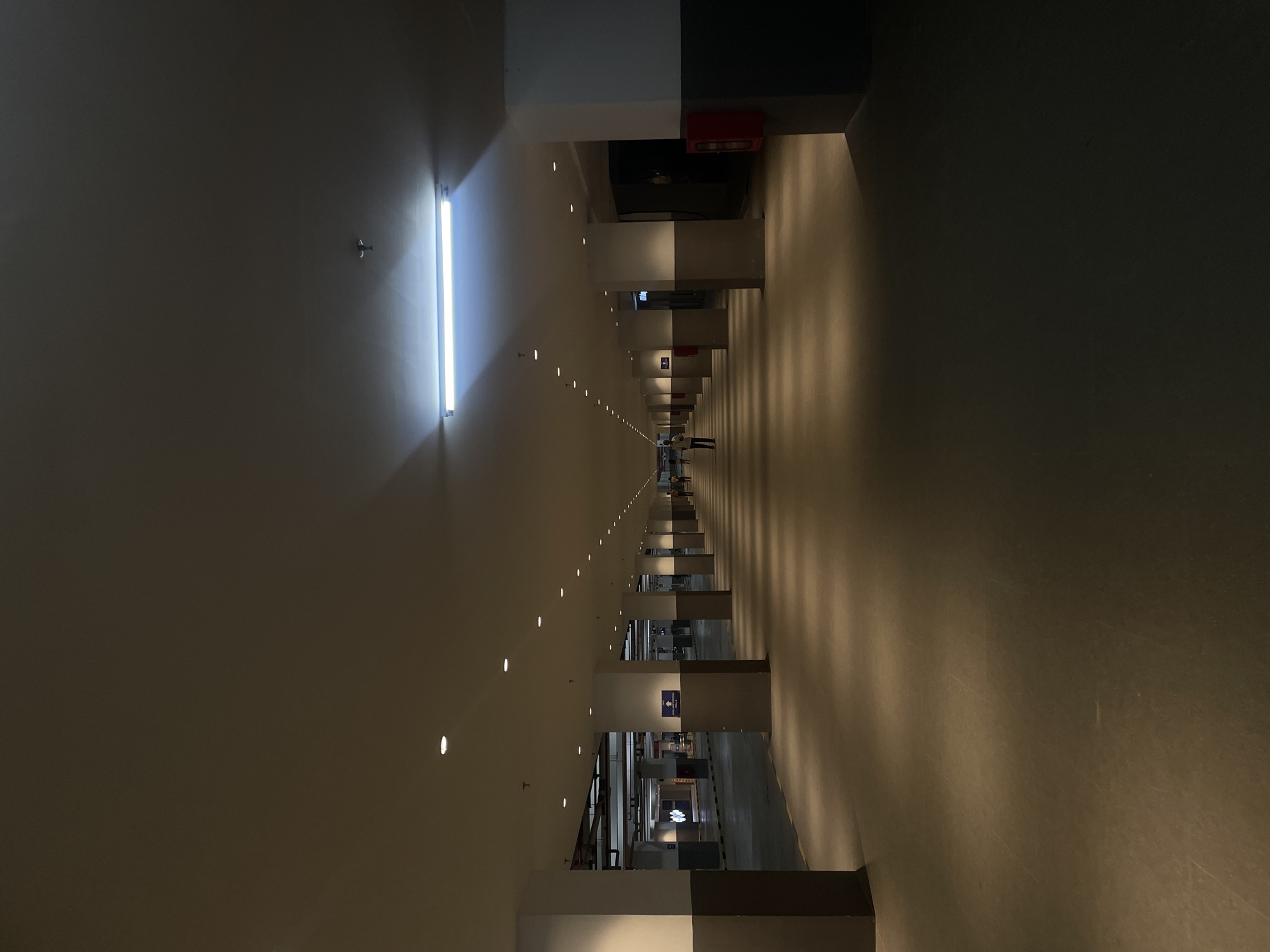 The image shows a long, indoor parking garage or hallway with a high ceiling. The space is illuminated by a long fluorescent light at the near end, and a series of smaller recessed lights stretching into the distance. The walls are light-colored, possibly beige or off-white, and the floor appears to be concrete. There are structural columns on both sides of the corridor, some painted with white and dark gray portions. In the distance, several people stand near the vanishing point of the perspective. Visible signage and indications suggest the location might be in Salembaran, Indonesia. A red fire extinguisher box is visible on a column near the top of the image. The perspective emphasizes the depth of the space.