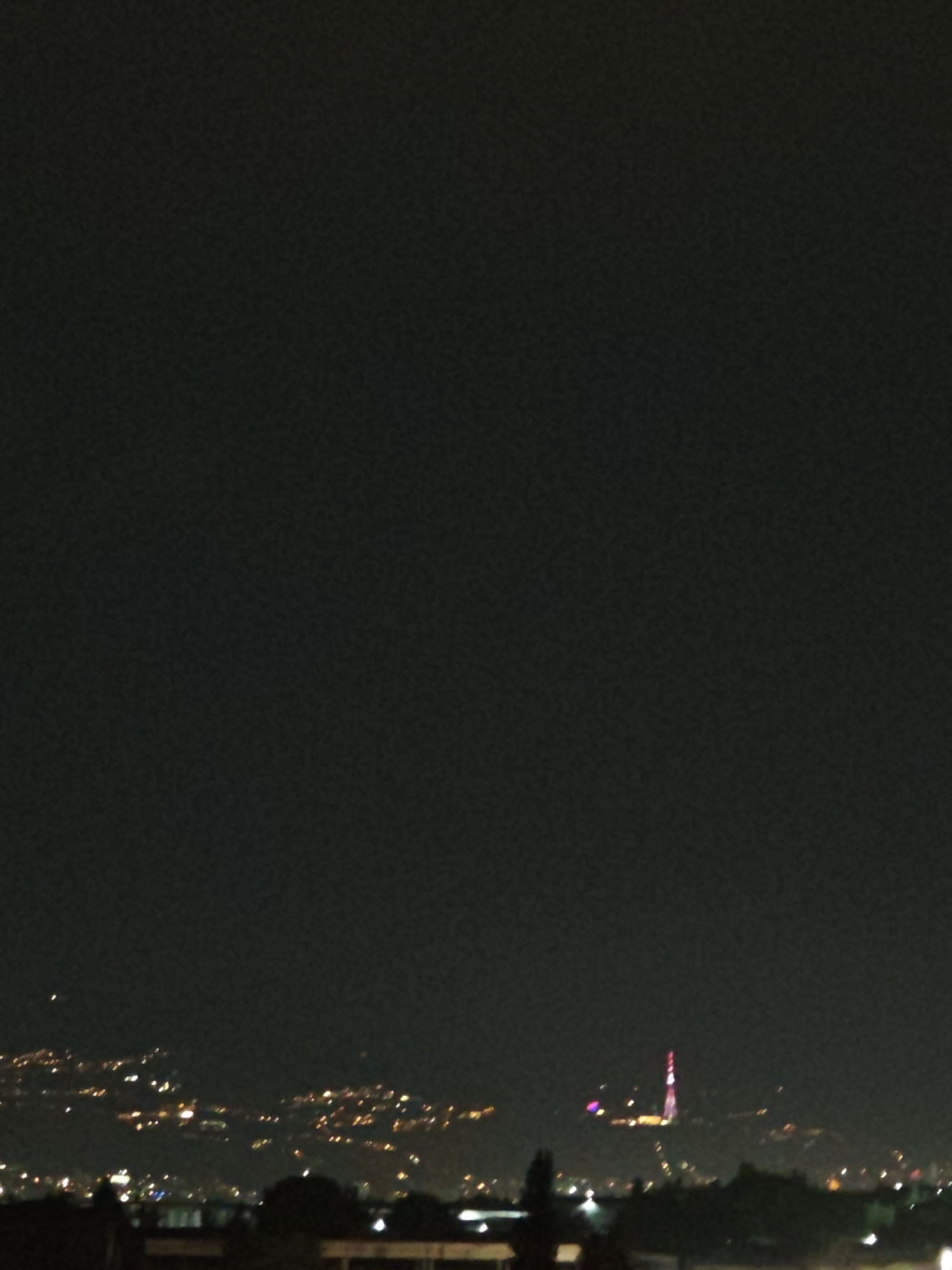 The image shows a night view of Tbilisi, Georgia, with the city lights spread across rolling hills. In the foreground, dark silhouettes of buildings and trees are visible. A prominent structure, likely the Tbilisi TV Tower, stands out in the mid-ground, illuminated with red and white lights. Numerous smaller lights indicate residential areas and streets spread throughout the cityscape. The sky is dark, suggesting it is nighttime.