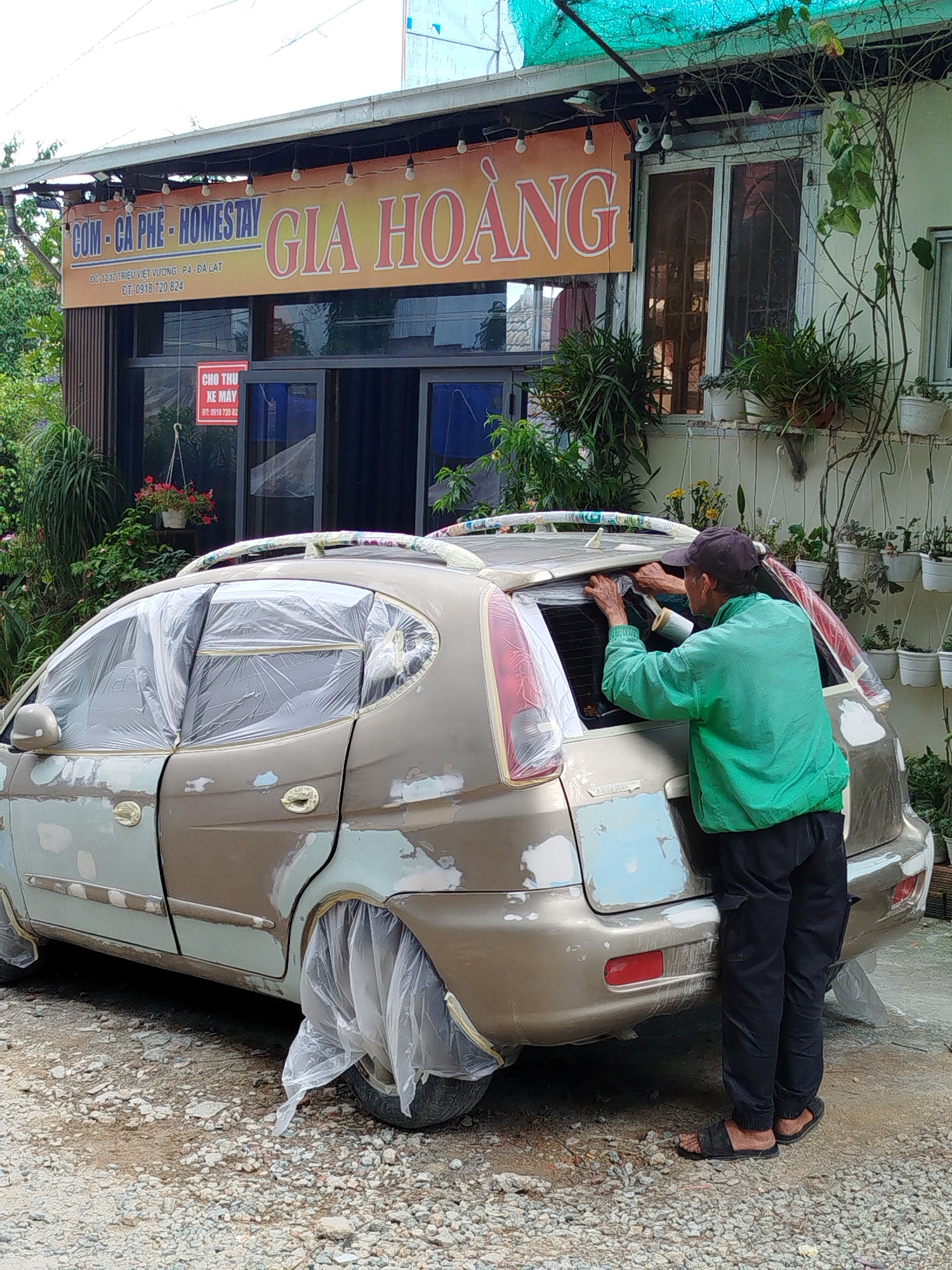 An elderly man in a black cap, green shirt, and black pants, is shown working on the back of a gold Daewoo Matiz parked on a gravel surface in front of the "COM-CA-PHE-HOMESTAY GIA HOANG" shop located at DC:12/12 TRIEU VIET VUONG-P4-DA LAT in the city of Da Lat, Vietnam. The car appears to be in the process of being repaired or repainted, with patches of putty on its body and clear plastic covering the windows and wheels. The shop has a sign in front of it and plants in pots along the building.