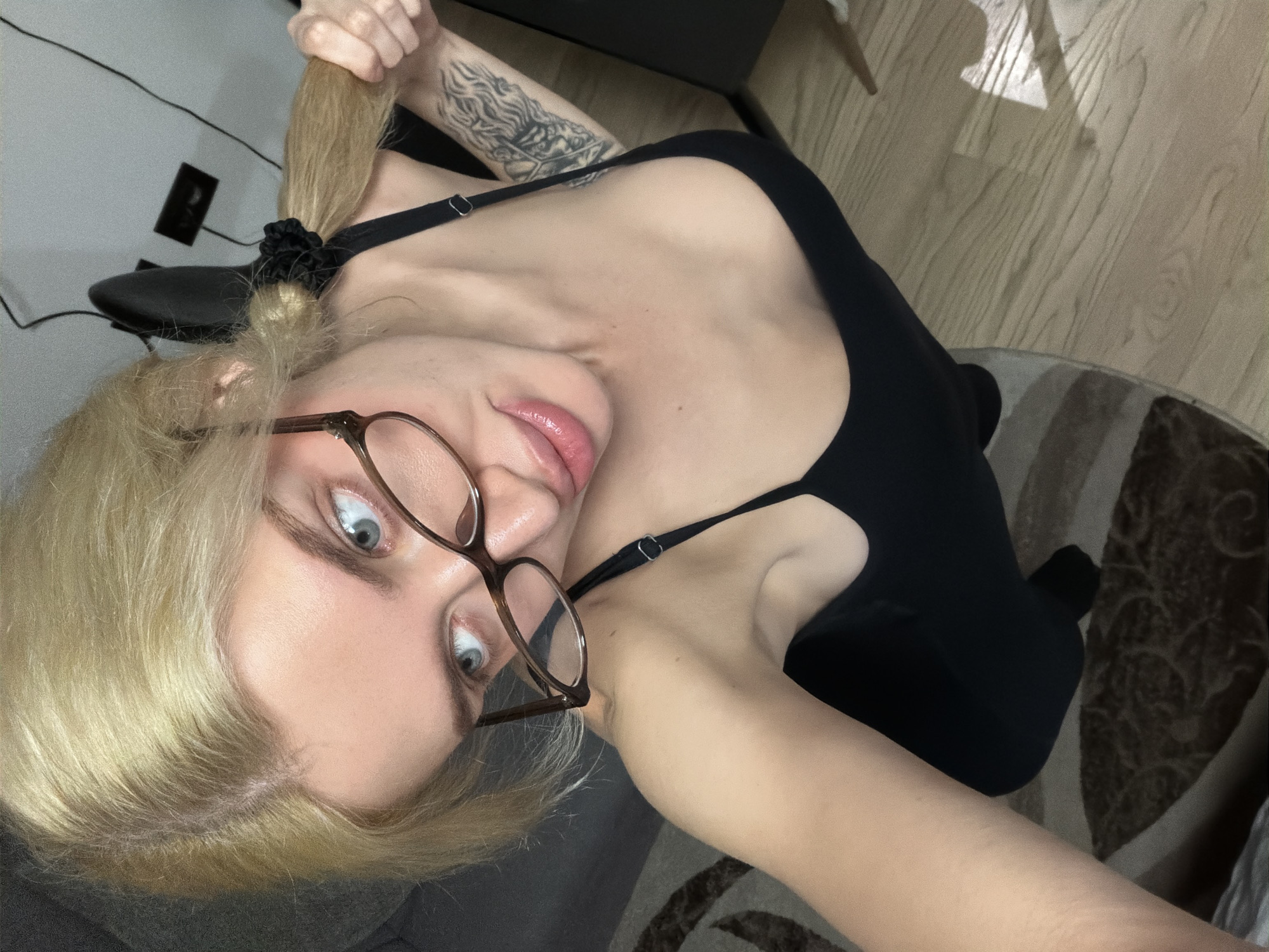 A young woman with light blonde hair, wearing glasses and a black tank top, is looking directly at the camera with wide eyes. Her hair is pulled back into a ponytail held by a black scrunchie. She is holding the end of her ponytail with her hand. A tattoo is visible on her left forearm. The background includes a wooden floor, a striped rug, and a black chair. The location appears to be indoors. No information indicates that this image was taken in Belgrade, Serbia.