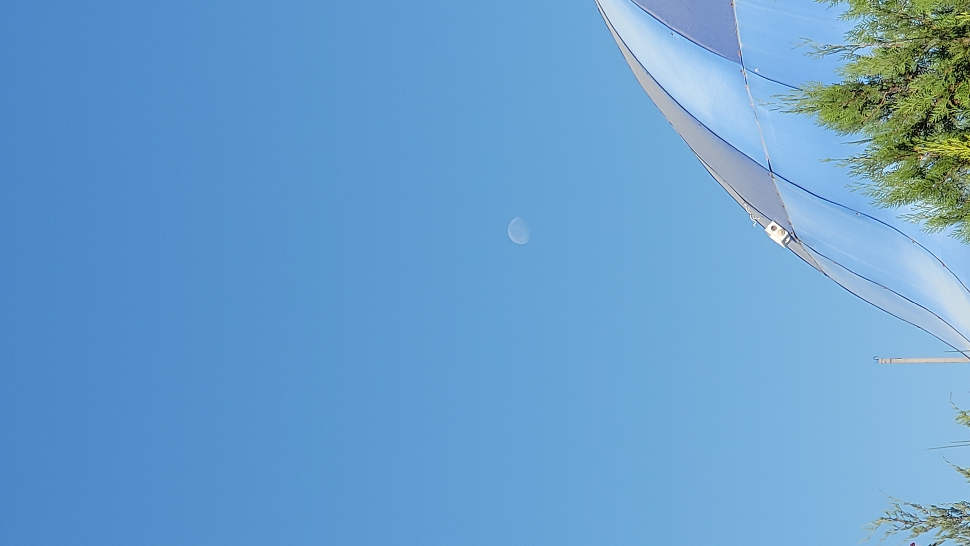 The image captures a clear blue sky with the moon visible in the upper middle section. In the lower right quadrant, the curved, blue and white striped surface of a structure, possibly part of a water slide or a dome, extends into the frame. Green coniferous trees are visible in the foreground, partially obscuring the base of the structure. A small white electronic device with a visible wire is attached to the curved surface of the structure. The scene appears to be outdoors in an open area. There are no people or animals in the image. The location is identified as Gulabad, Uzbekistan.