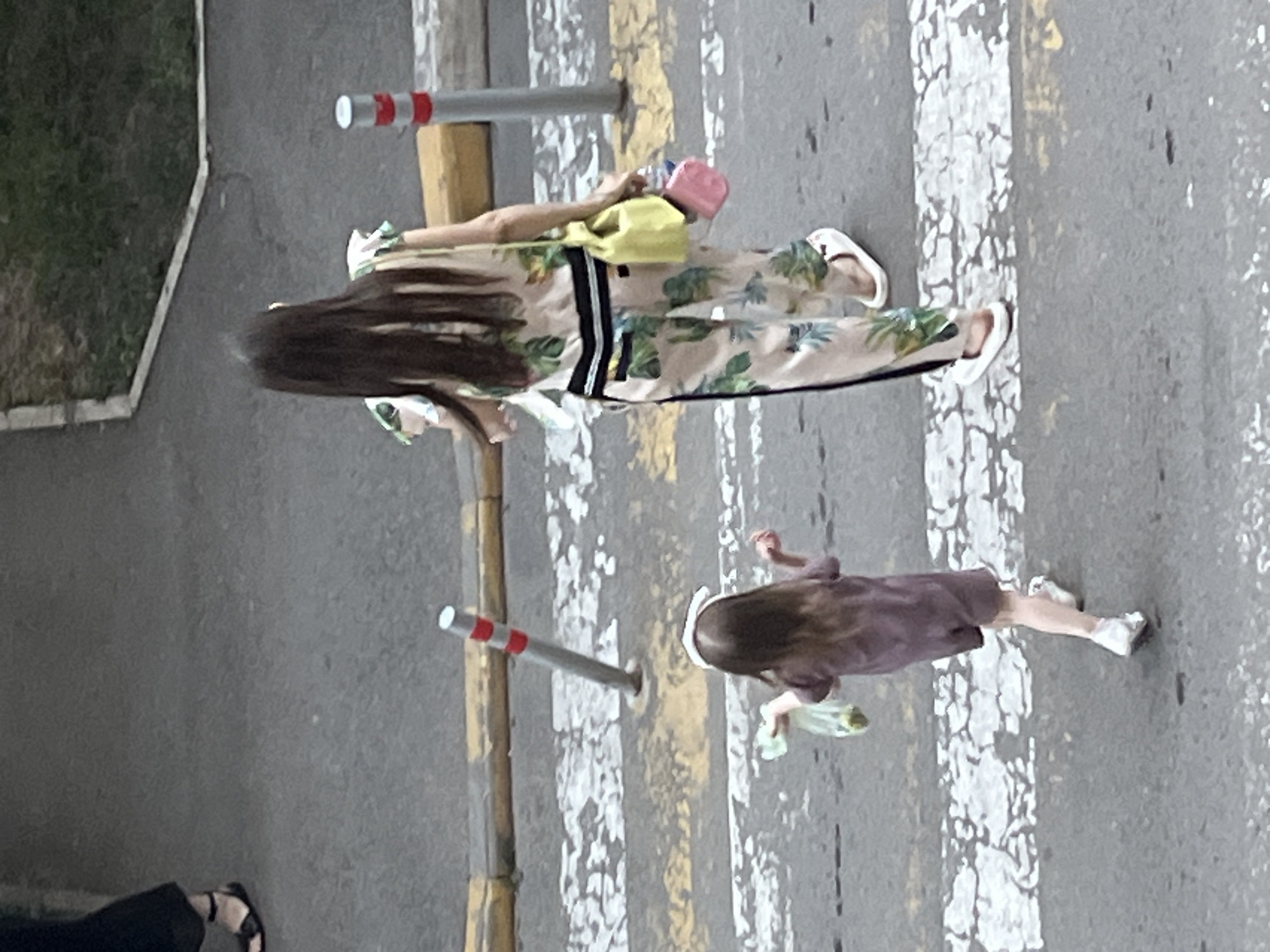 The image captures an aerial view of a woman and a young child crossing a pedestrian street in Krasnodar, Russia. The woman, dressed in a tropical-print jumpsuit and white slides, carries a yellow purse and a pink clutch. The child, wearing a plum-colored dress and white sneakers, holds a clear plastic bag in their left hand. Both are walking from left to right across a zebra crossing marked with white stripes. The street is paved with asphalt, with a yellow and gray curb visible in the background. Two red and white striped bollards are positioned along the curb, one leaning slightly to the left. A patch of grass and a concrete edge are visible beyond the curb. The overall lighting suggests it is daytime, possibly overcast given the soft shadows.